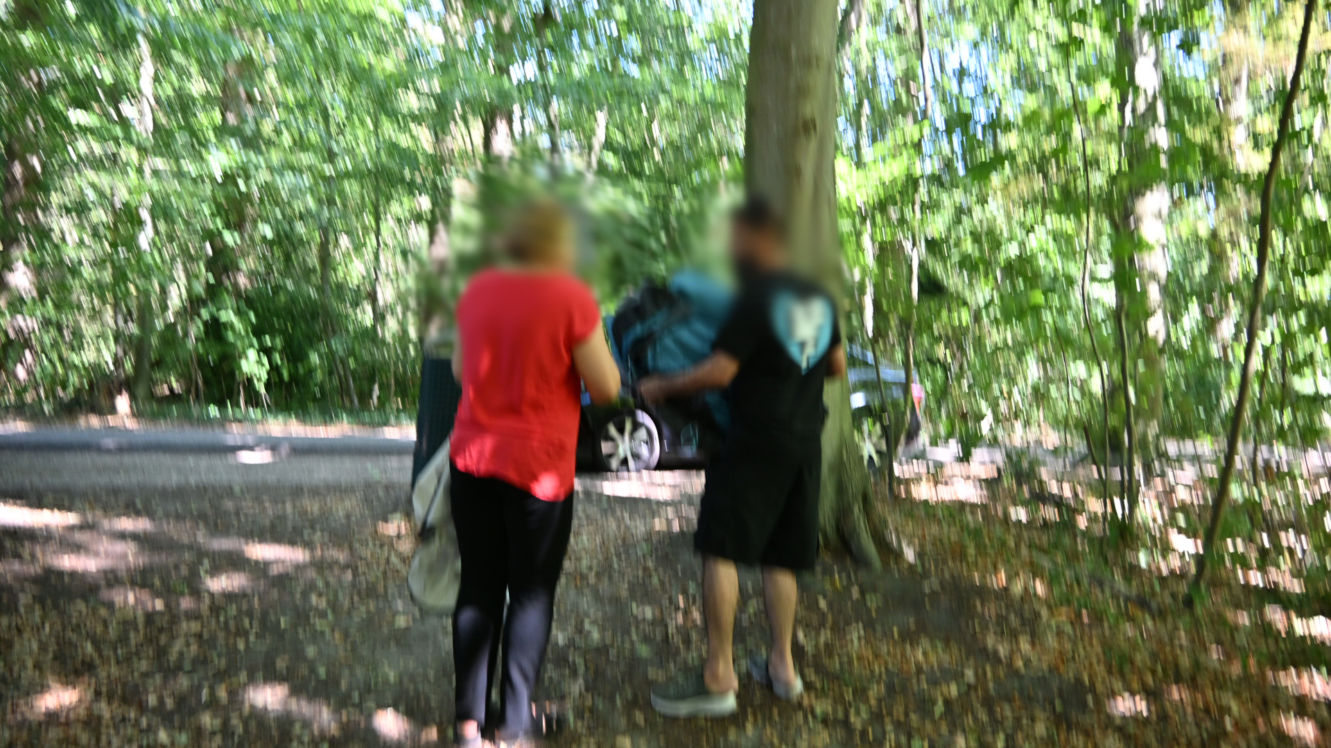 Twee personen staan op een bospad bij een boom, met een geparkeerde auto op de achtergrond
