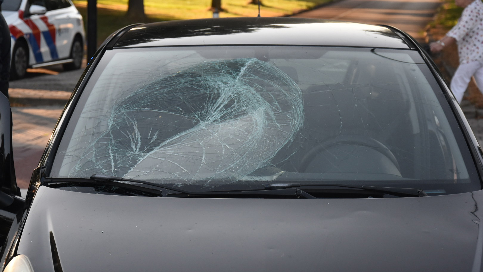 Voorruit van een auto met grote barst en spinwebscheuren na een aanrijding, met politieauto op de achtergrond