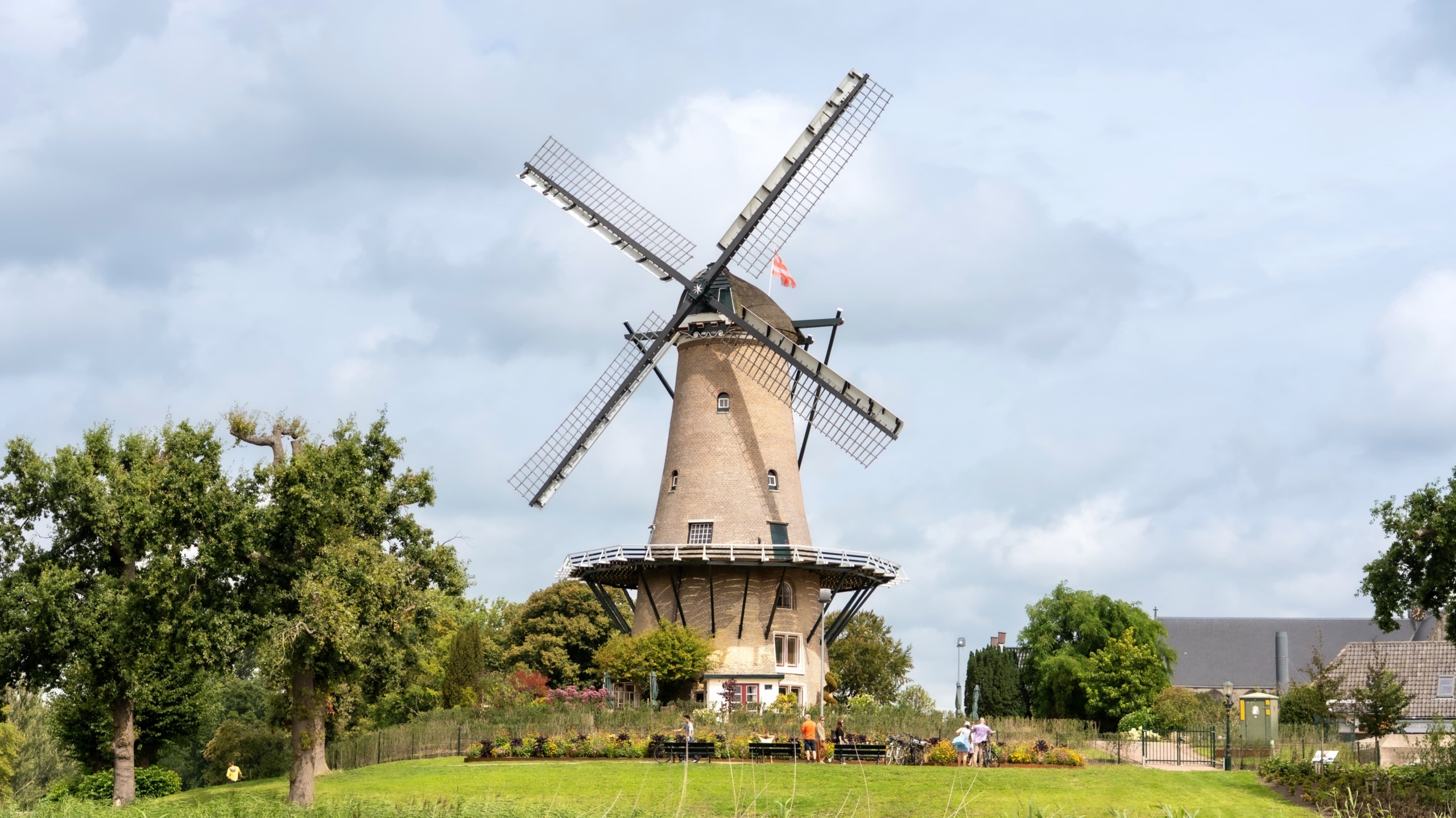 Bakstenen windmolen met grote wieken op een groene heuvel, met bomen en een paar mensen onder een bewolkte lucht
