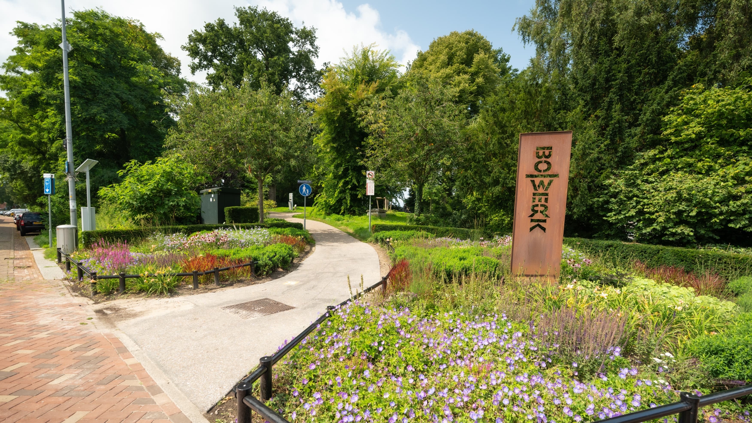 Ingang van het Bosje van Weck met slingerend pad, bloemenperk en hoge roestkleurige zuil met tekst