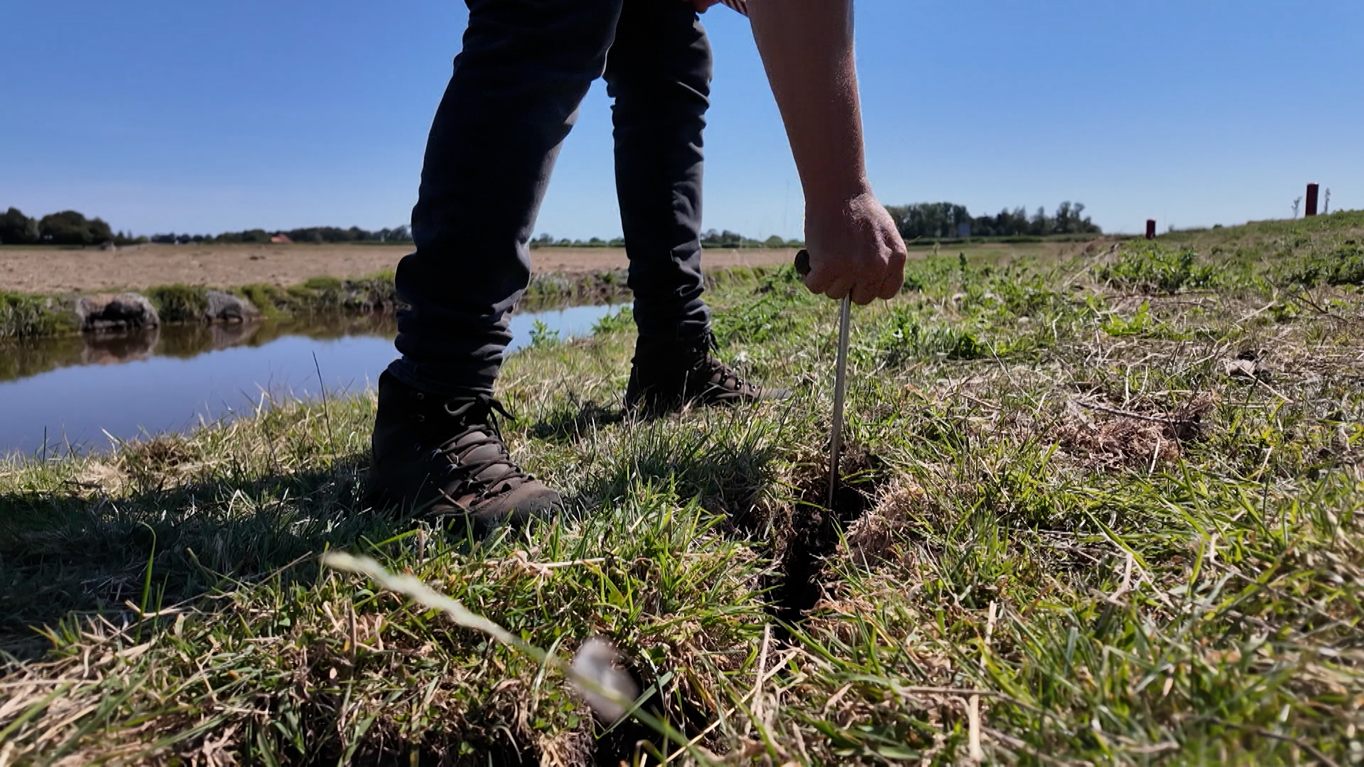 Persoon steekt een metalen pen in de grasdijk naast een sloot, met laarzen en akkerland op de achtergrond