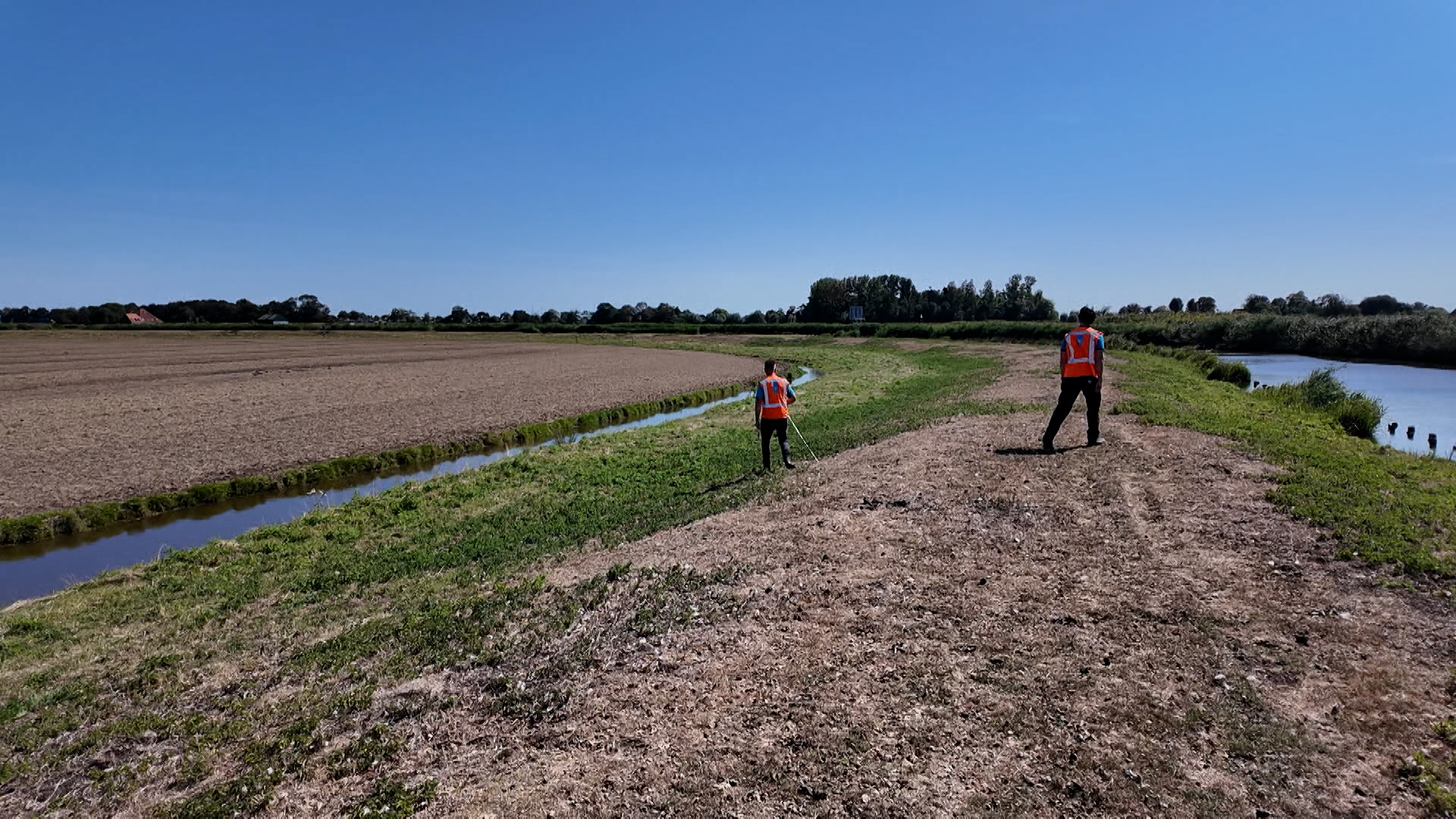 Twee mensen in oranje veiligheidsvesten lopen op een dijkpad langs een sloot in open polderlandschap