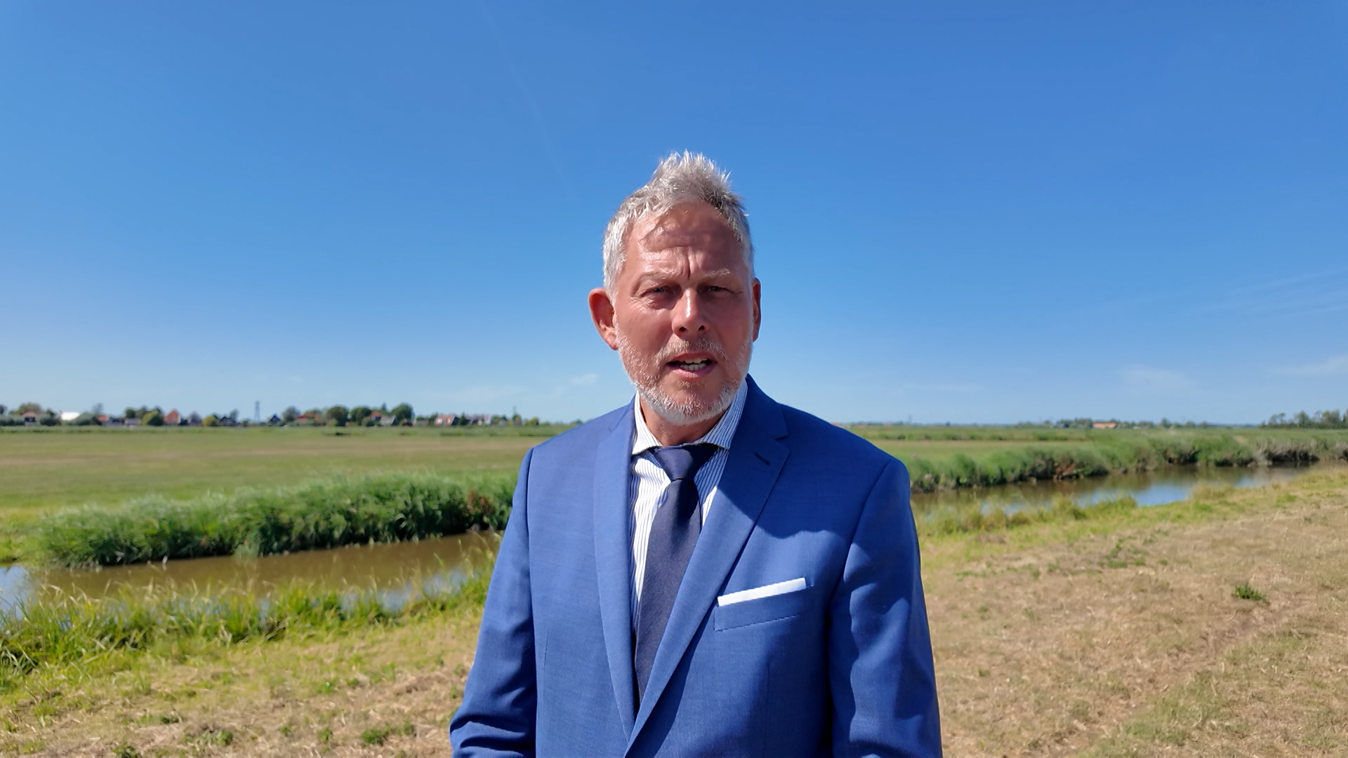 Man in blauw pak en stropdas staat buiten bij een sloot in een groen polderlandschap onder blauwe lucht
