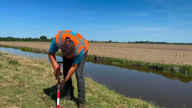 Man in oranje veiligheidsvest plaatst een meetstok in de grond langs een sloot in een open polderlandschap