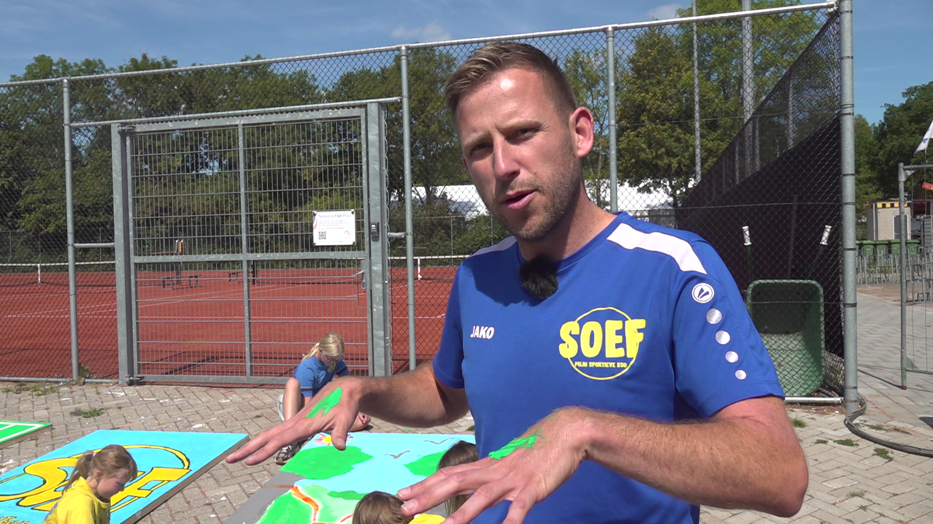 Man in blauw SOEF-shirt met groene verf op handen spreekt bij tennisbaan terwijl kinderen op panelen schilderen