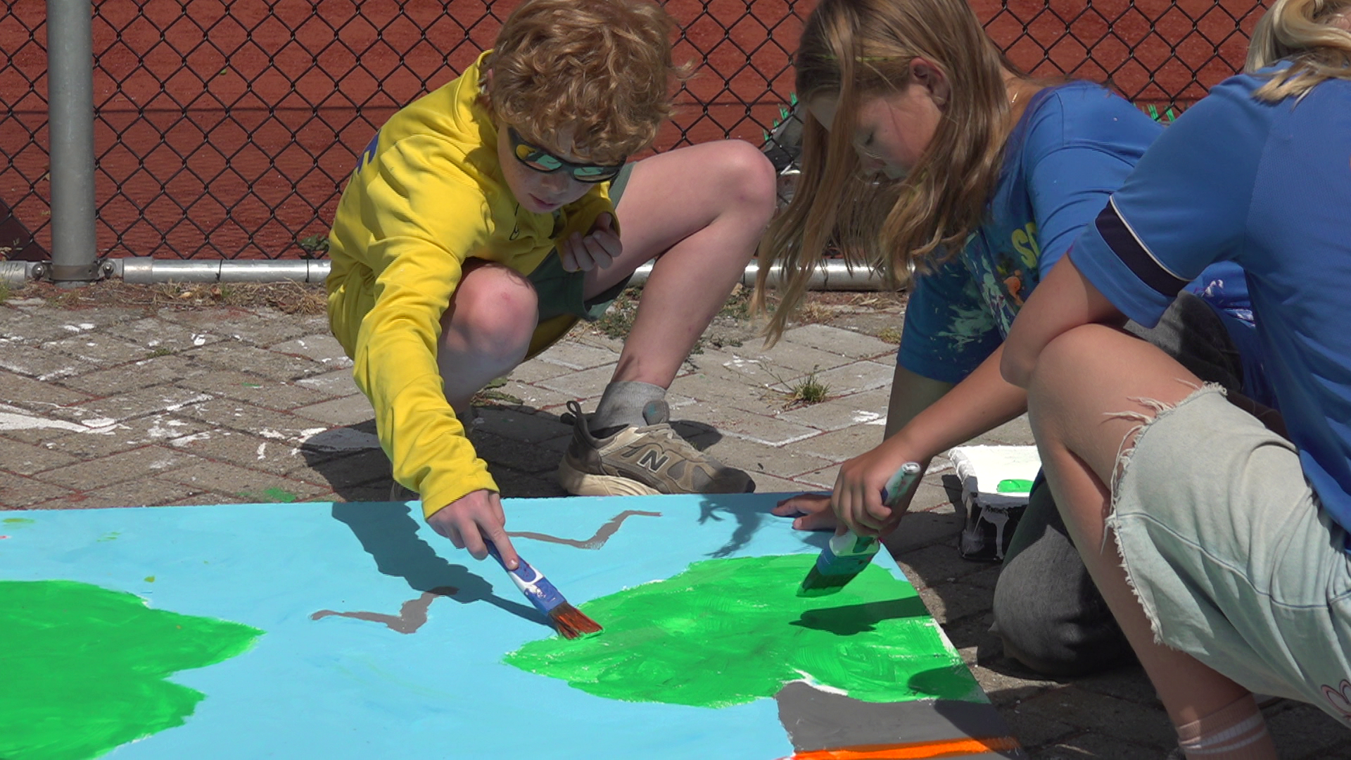 Kinderen schilderen samen een grote blauwgroene tekening op de stoep met kwasten, bij een hek op een sportveld