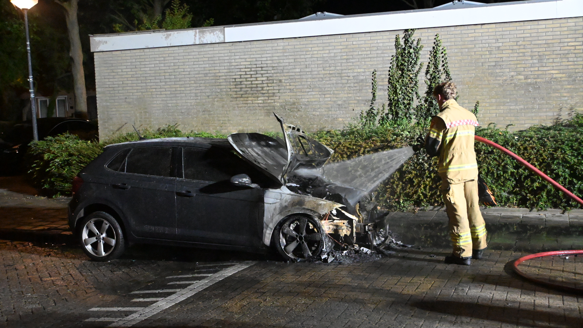 Brandweerman blust brandende auto met zwaar beschadigde voorkant, met open motorkap en rook in het donker