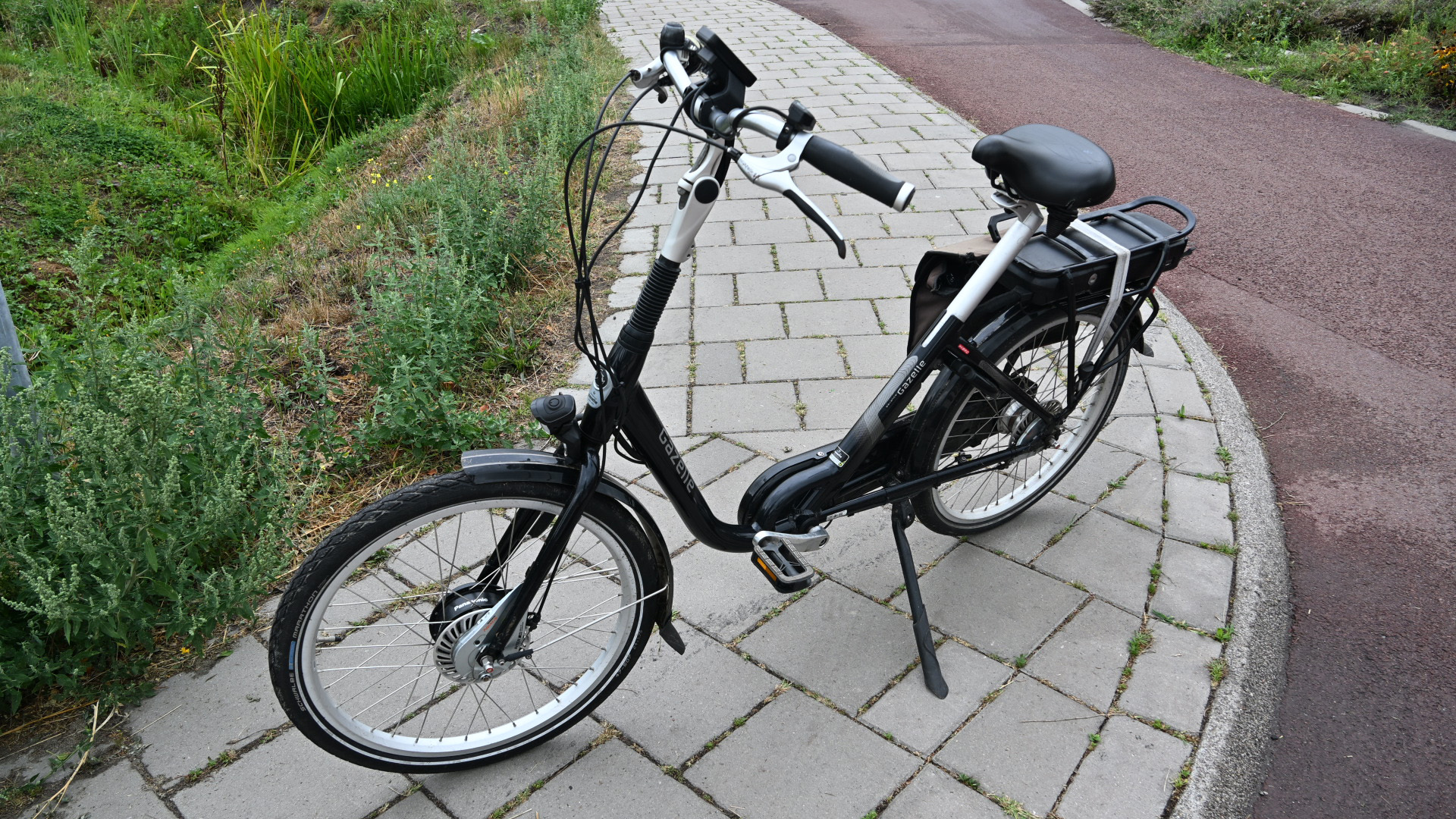 Zwarte elektrische stadsfiets met lage instap en accu op de bagagedrager, geparkeerd op een stoep langs een fietspad