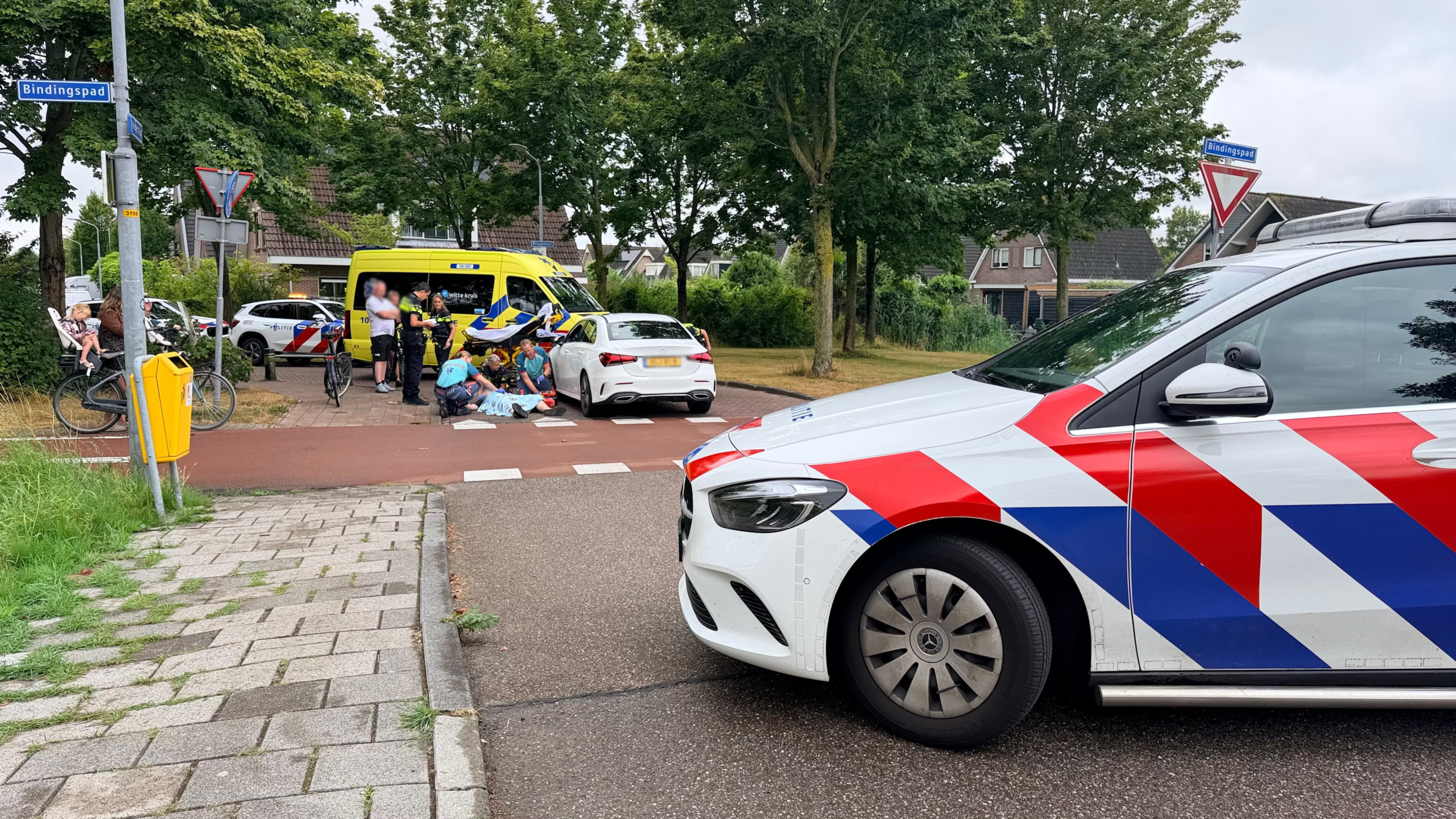 Politieauto op de voorgrond terwijl hulpdiensten een slachtoffer behandelen bij een ongeval met witte auto en ambulance