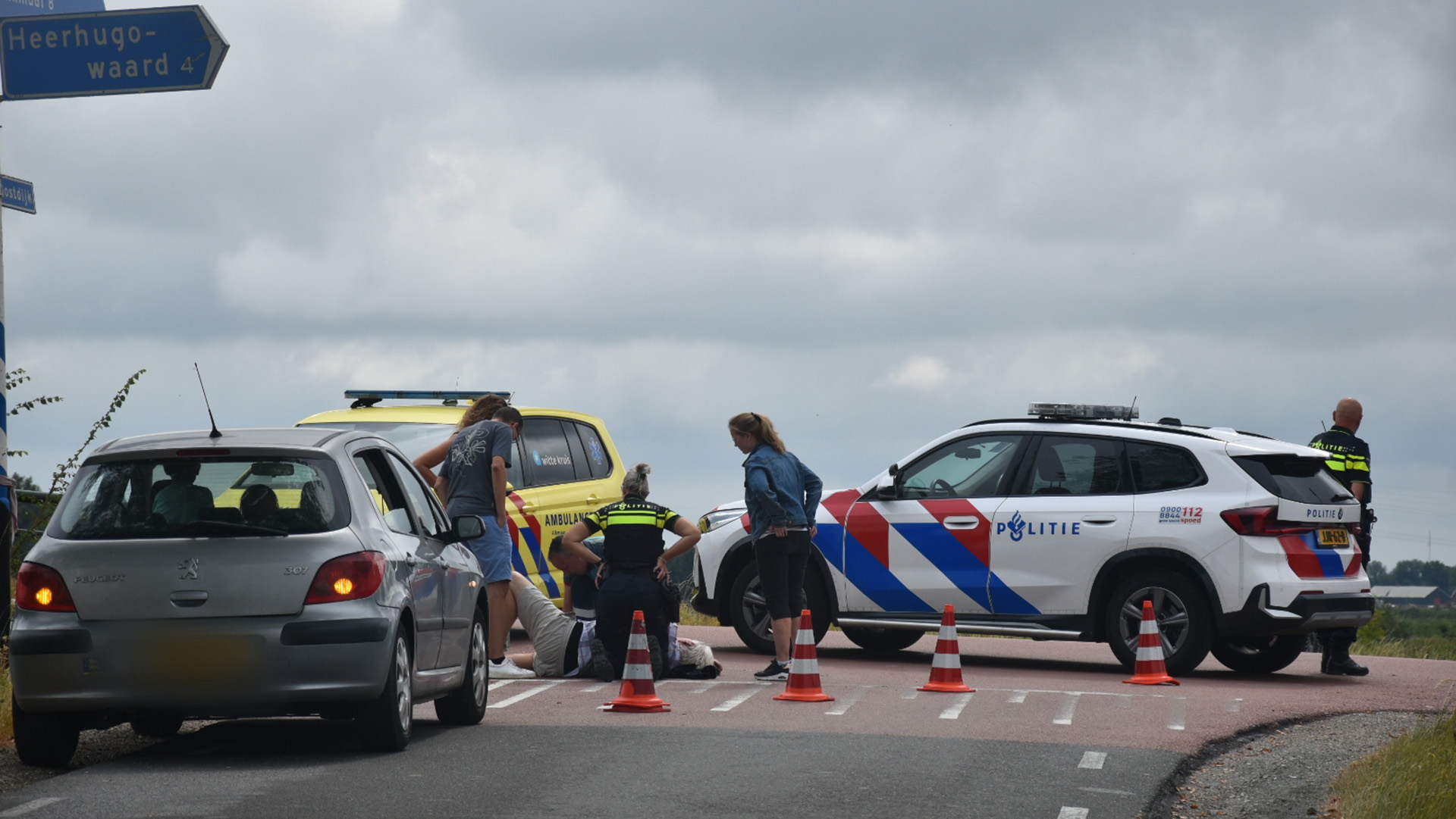 Politie en ambulance bij verkeersongeval; persoon ligt op de weg naast auto’s en pionnen, richtingbord “Heerhugowaard”