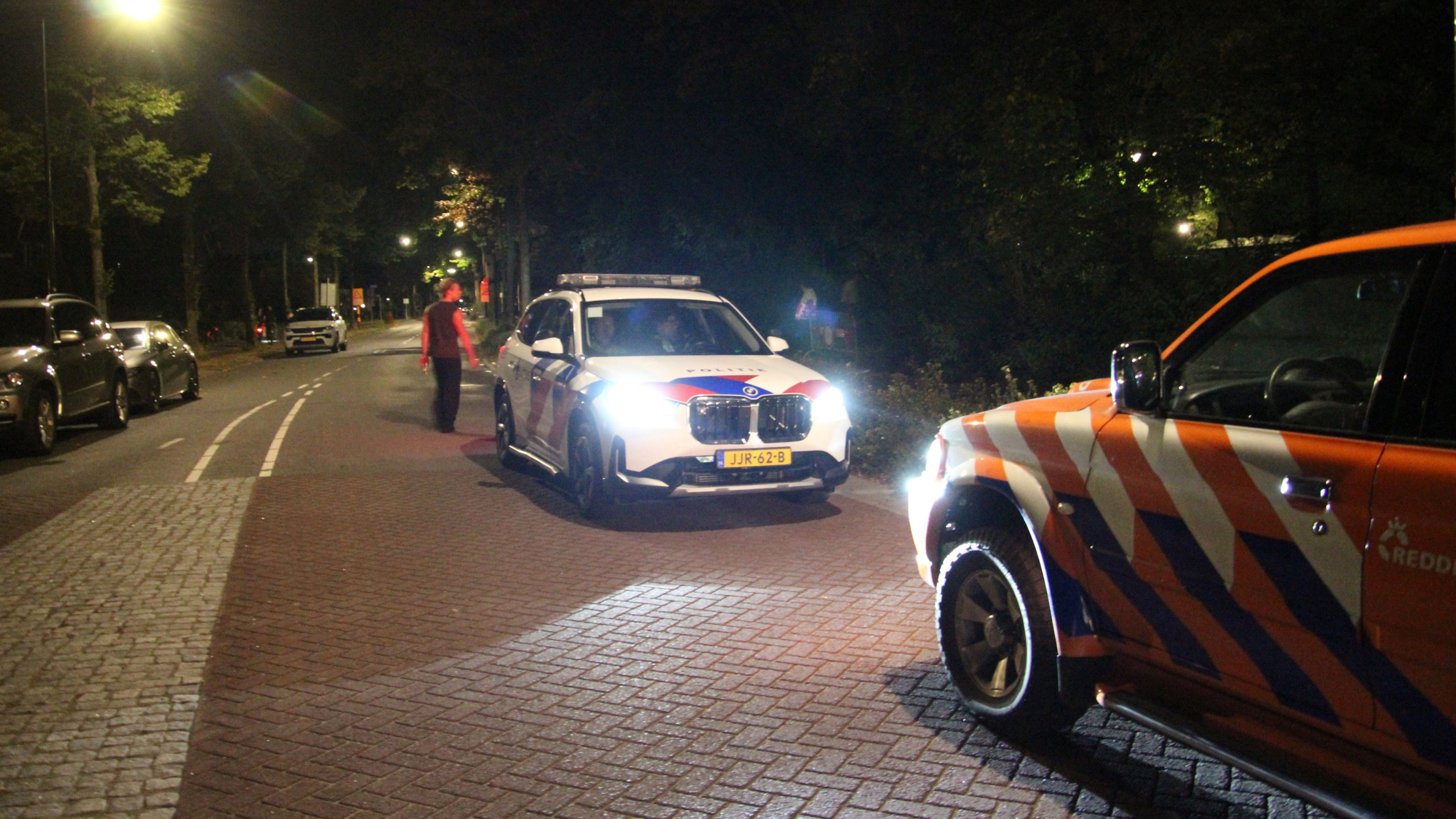 Politieauto met felle koplampen op een donkere straat, met een persoon lopend en een rood-wit gestreepte wagen op de voorgrond