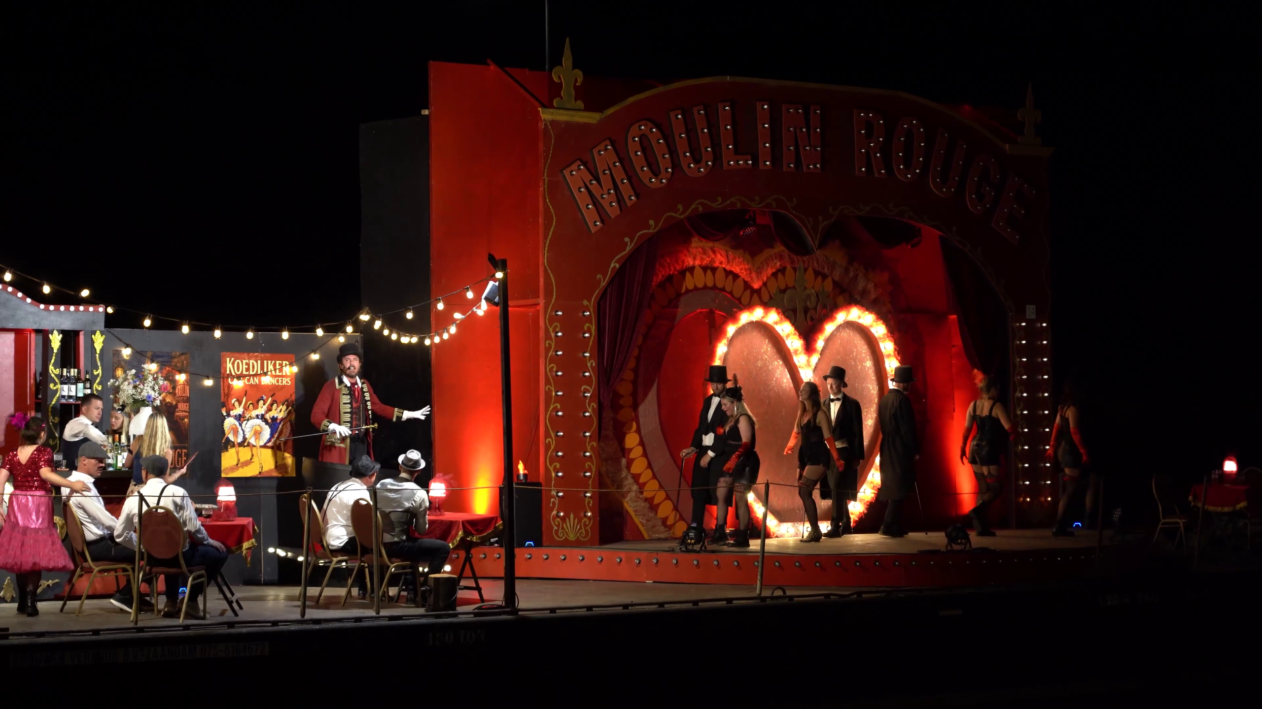 Avondvoorstelling in een rode Moulin Rouge-achtige cabaretzaal met dansers op het podium en publiek aan tafeltjes
