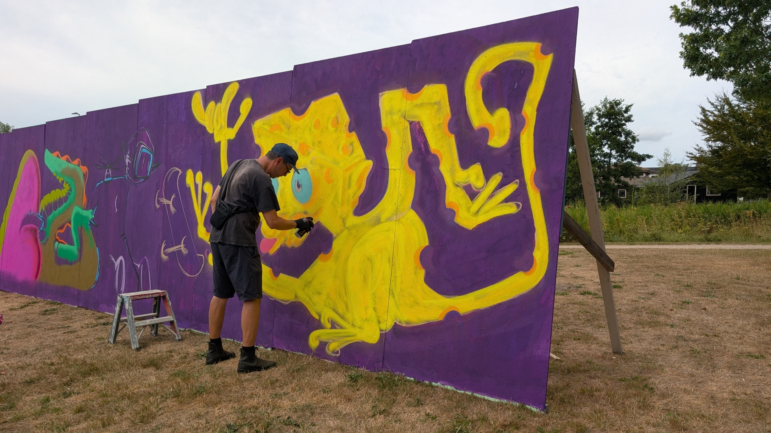 Man spuit graffiti op een paarse muur met een grote gele hagedis, met een krukje ervoor op een grasveld