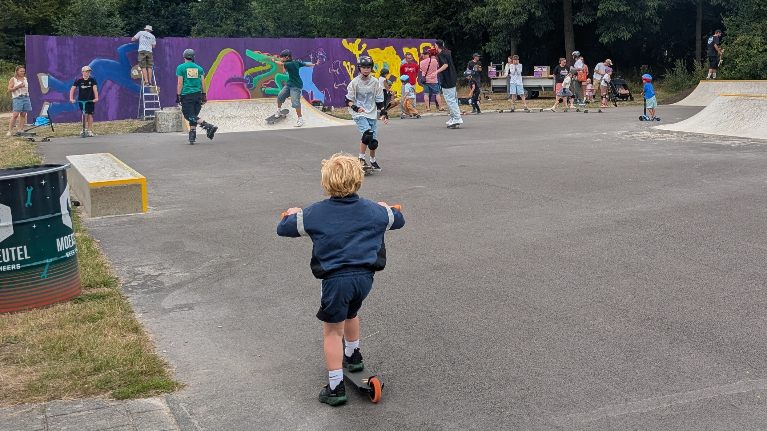 Kind op een step in een skatepark, met skaters en toeschouwers voor een muur met kleurrijke graffiti