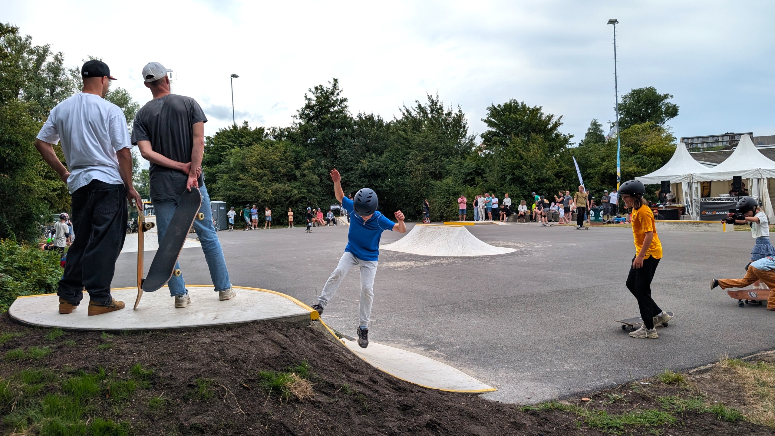 Skaters in een skatepark; een kind met helm springt van een ramp terwijl anderen toekijken en oefenen