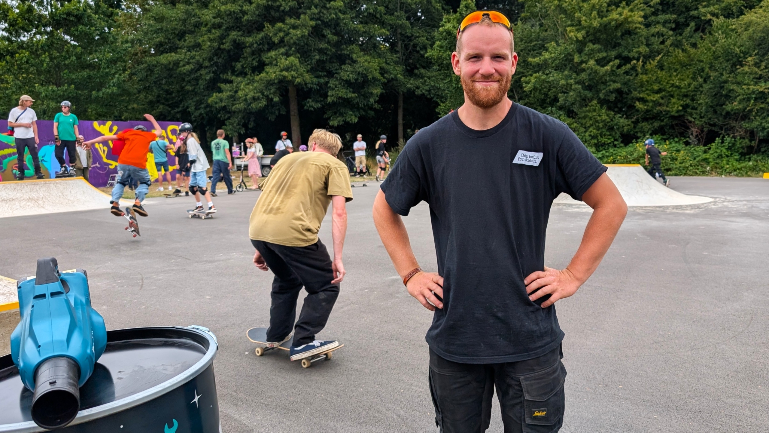 Man met zonnebril op het hoofd staat met handen in de zij bij een skatepark, terwijl skaters op de achtergrond tricks doen