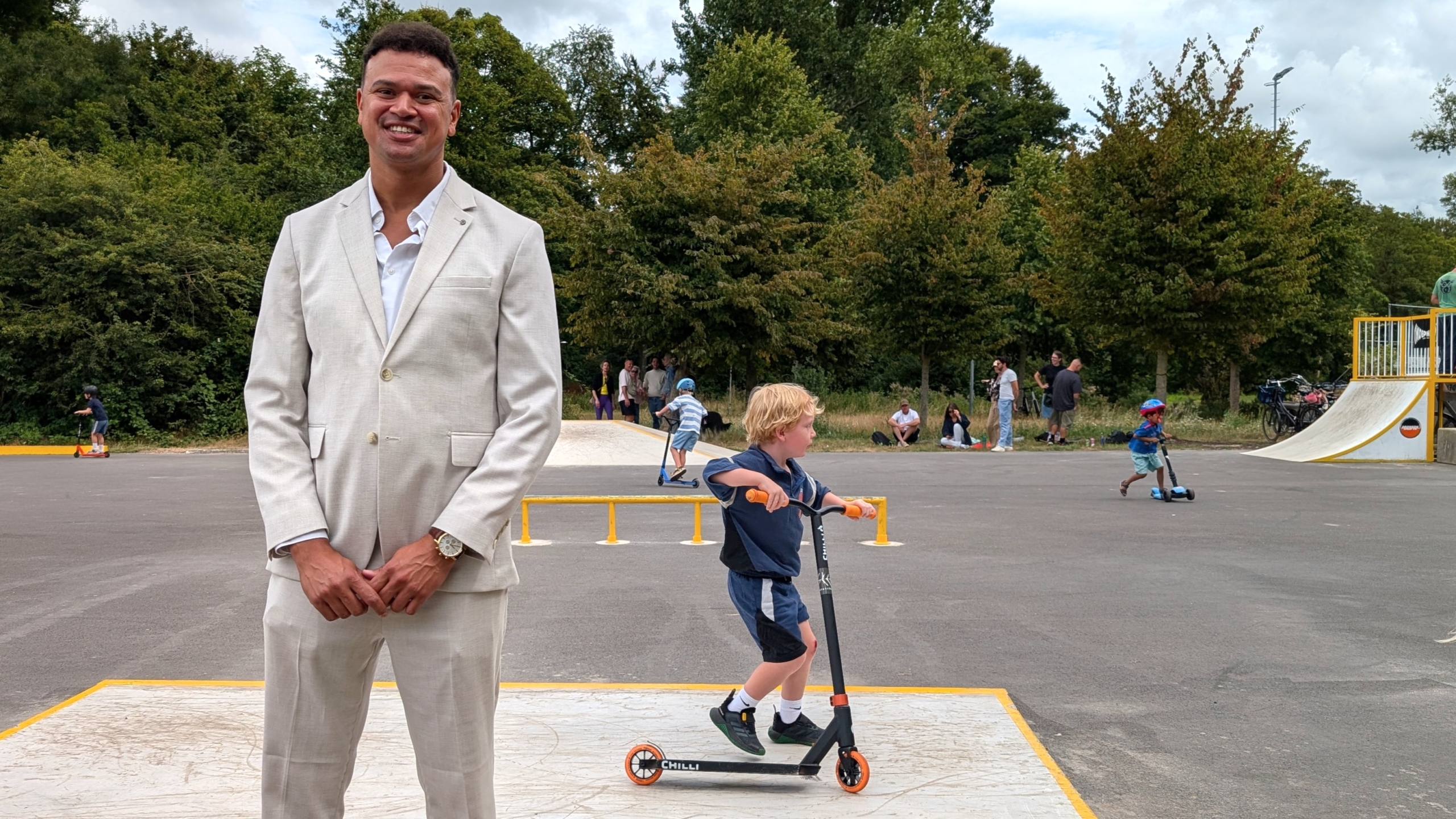 Man in licht pak glimlacht op een skatepark, terwijl kinderen op steps rijden en een ramp op de achtergrond staat