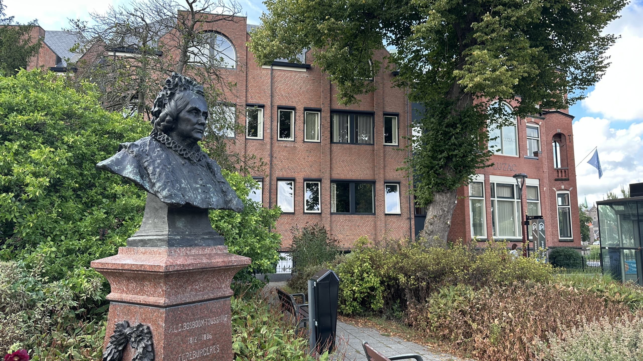 Bronzen buste op stenen sokkel in een groene tuin, met op de achtergrond een bakstenen gebouw met UZAC-logo