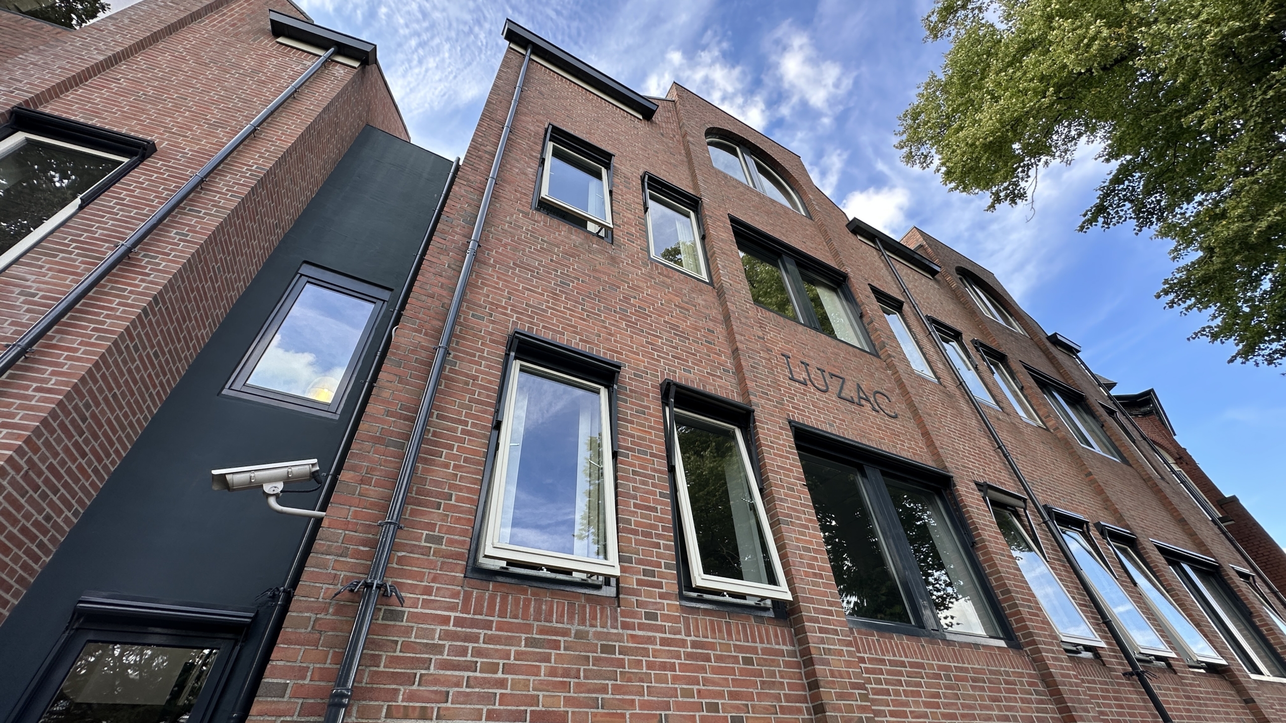 Opwaartse blik op bakstenen gebouw met zwarte kozijnen en het opschrift “LUZAC”, tegen blauwe lucht en bomen