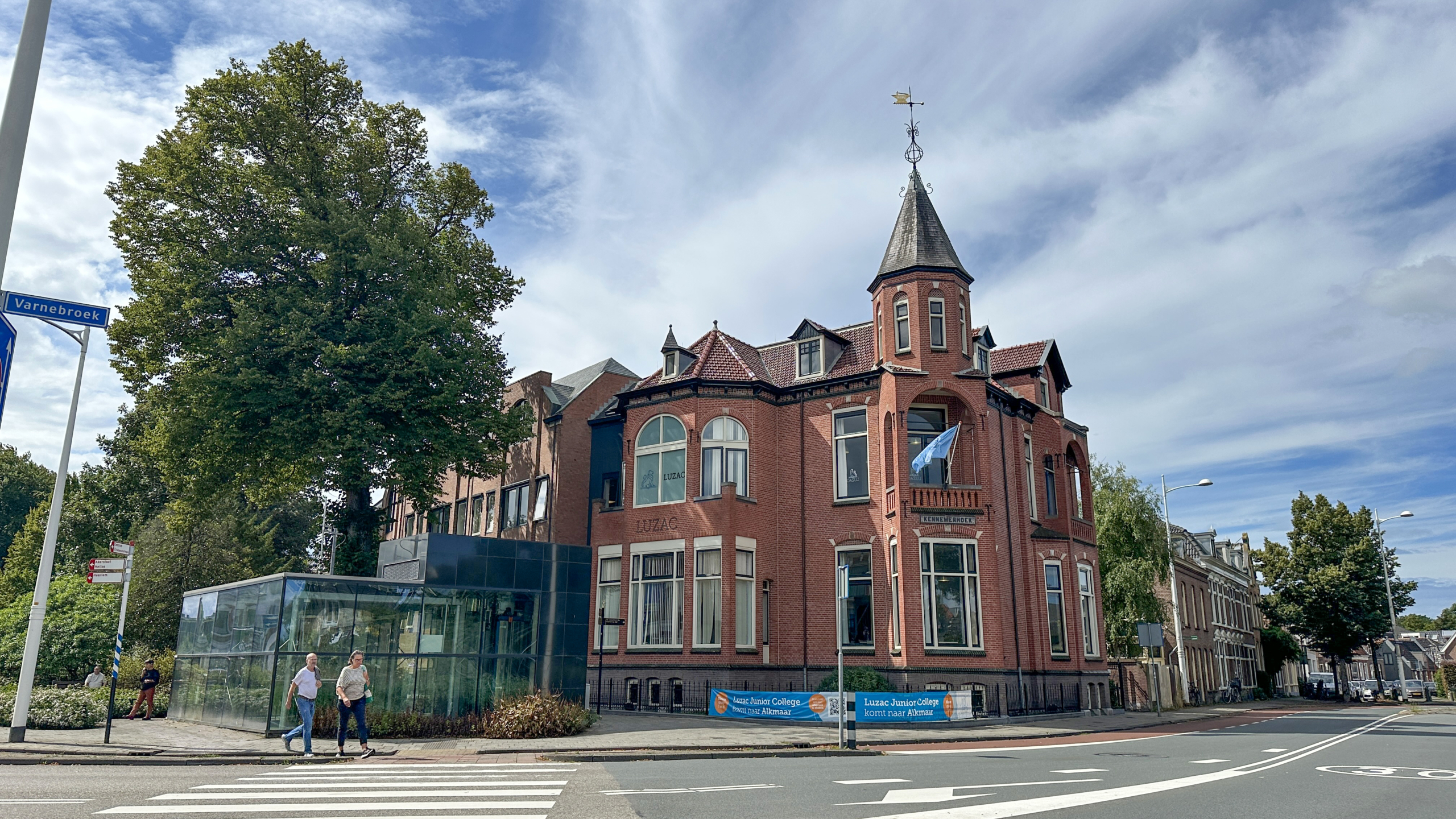 Rood bakstenen gebouw met torentje en windvaan naast glazen aanbouw, met grote boom en passerende voetgangers