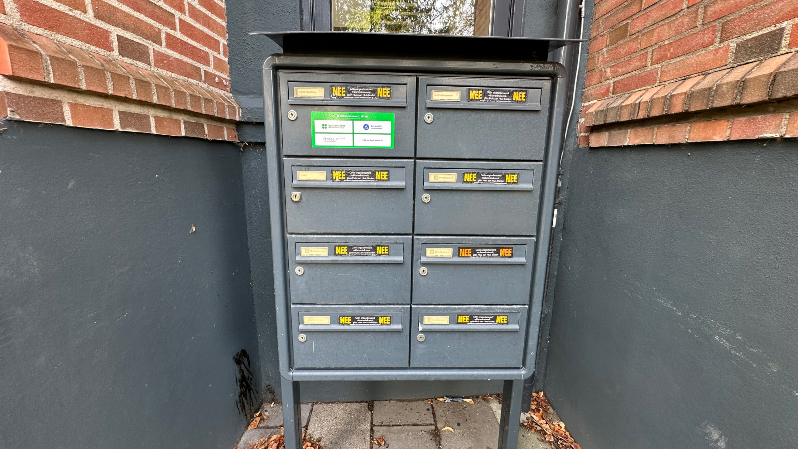 Grijze brievenbuskast met acht postvakken en “NEE”-stickers, tegen een bakstenen gevel bij een portiek