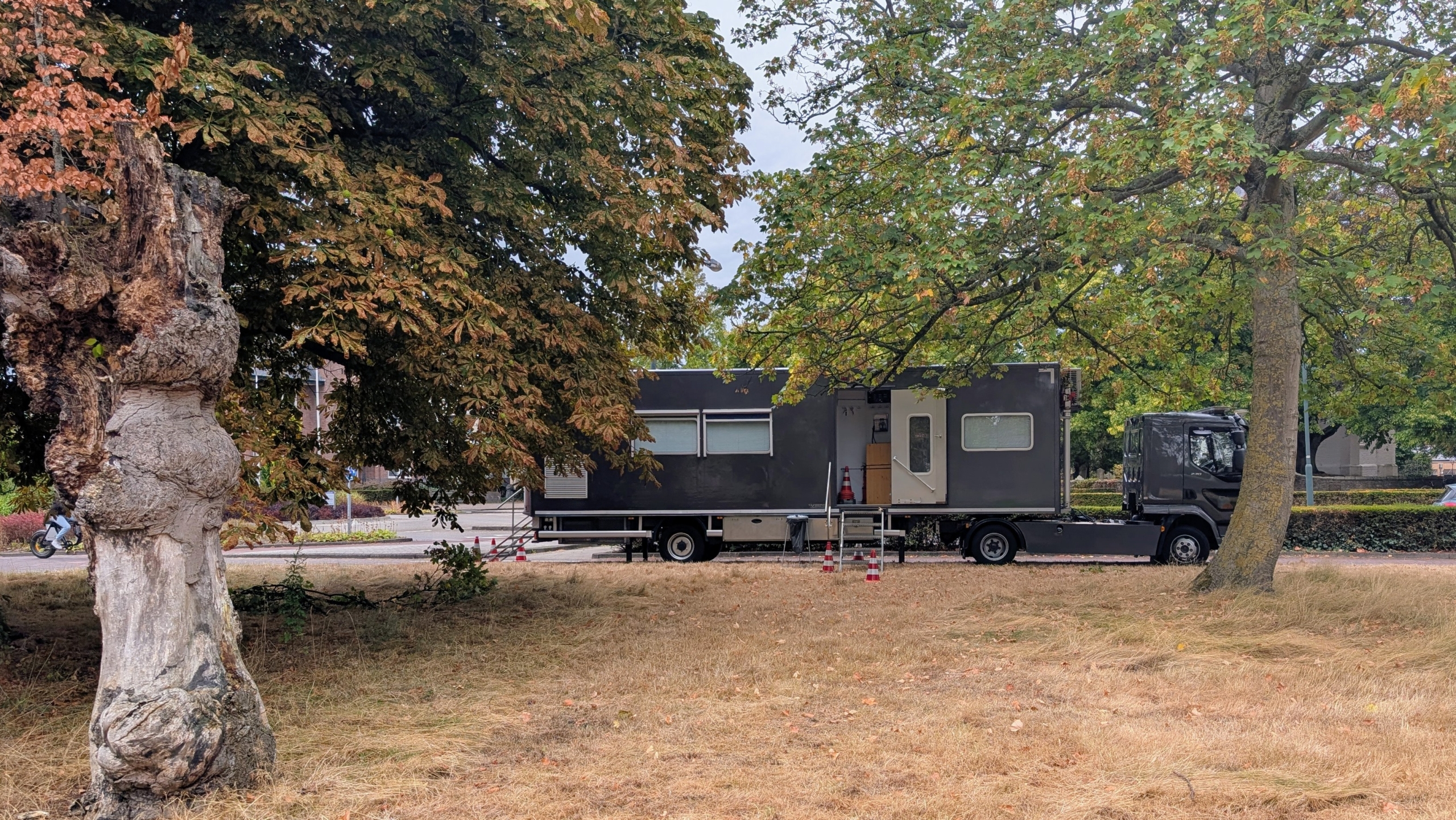 Zwarte vrachtwagen met mobiele unit geparkeerd onder grote bomen op een grasveld, met pionnen en trap bij de deur