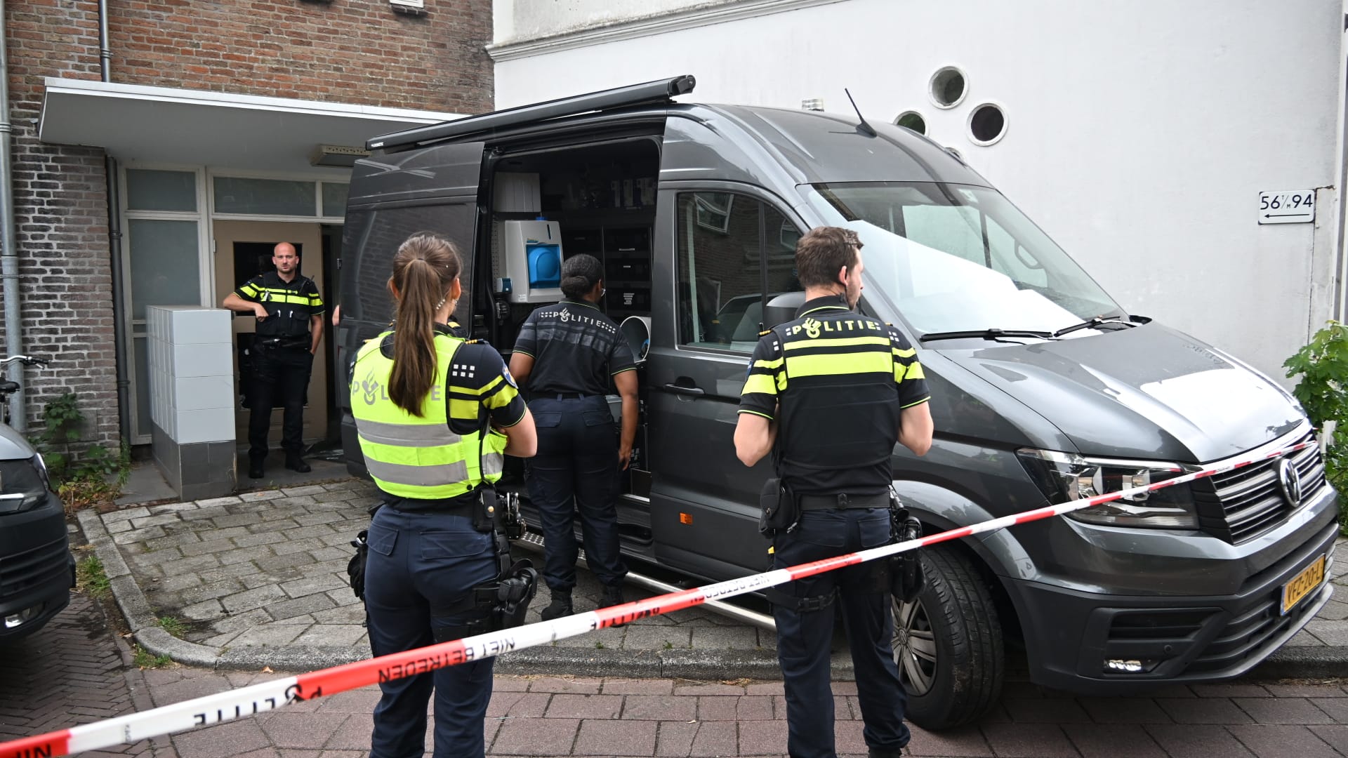 Politieagenten bij een donkergrijze politiebus voor een gebouw, met afzetlint op de voorgrond