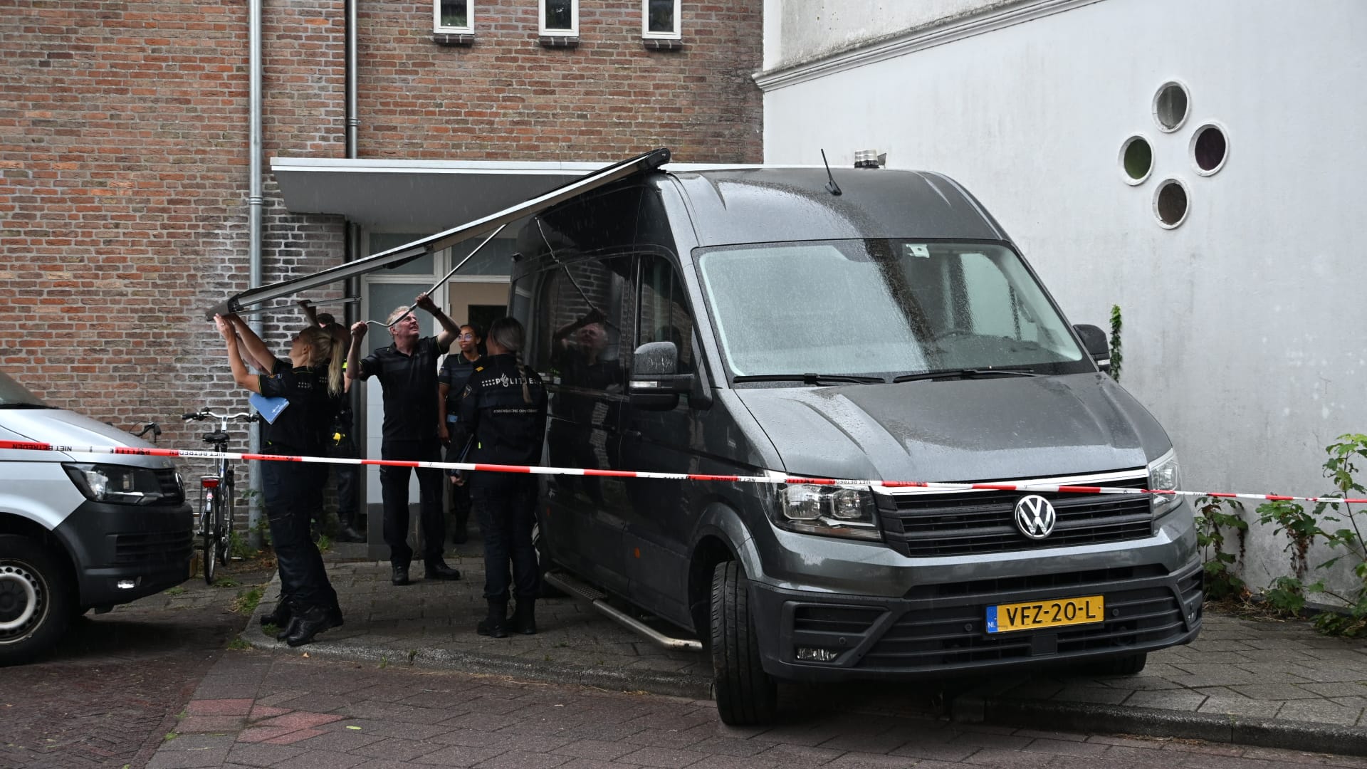 Agenten bij een afgezet gebied tillen een grote plaat van een grijze bestelbus naast een gebouw op