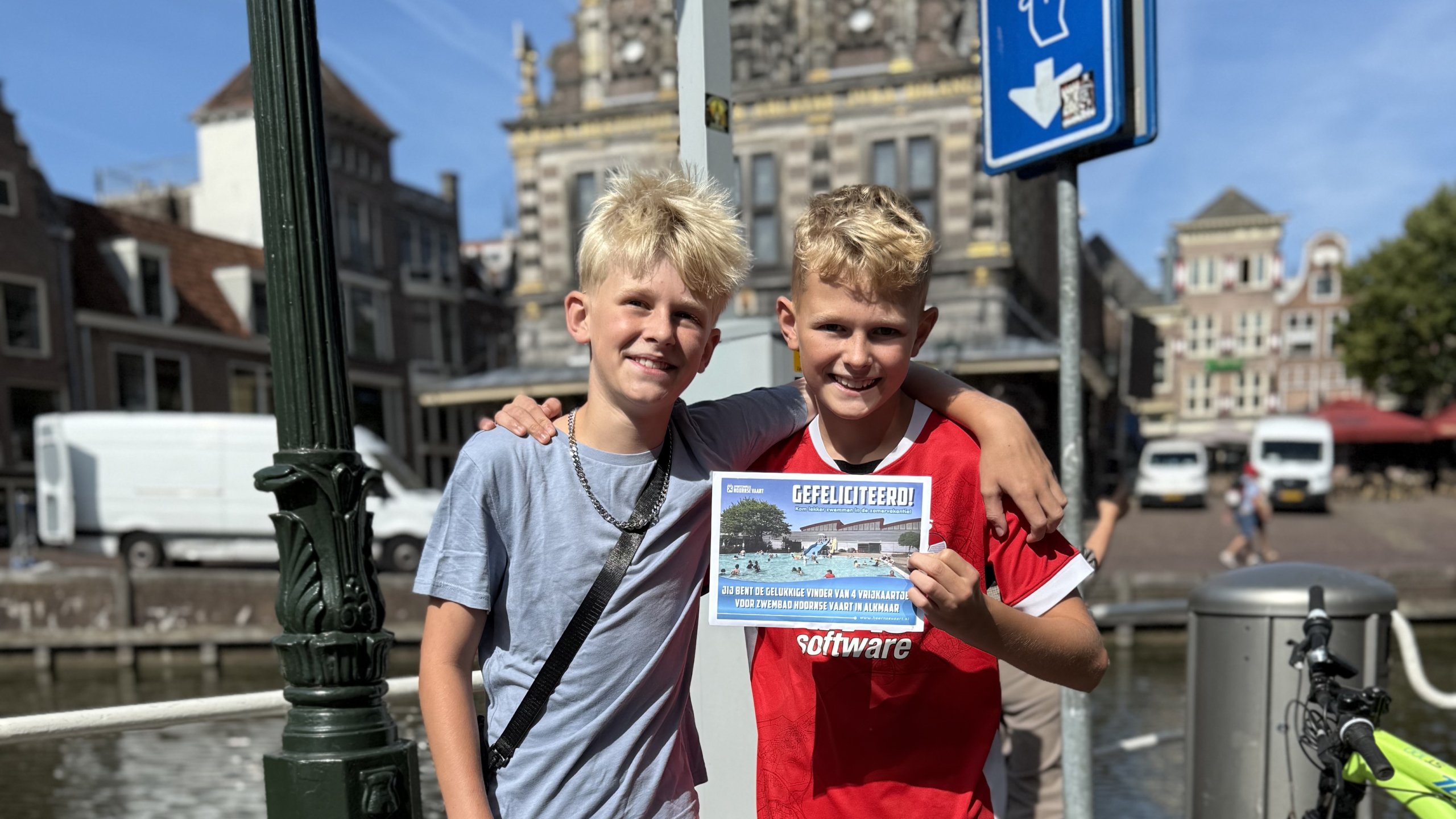 Twee jongens poseren op een brug; één houdt een flyer met de tekst “GEFELICITEERD!” vast, met stad op de achtergrond