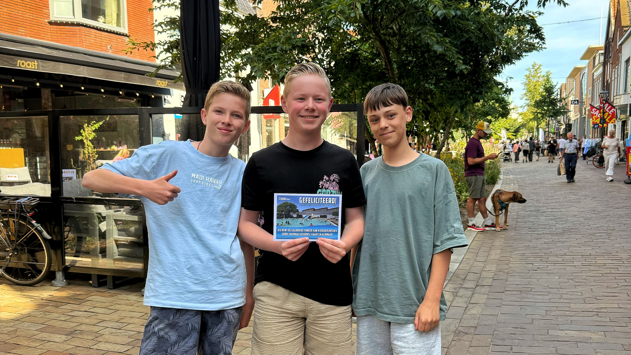 Drie jongens op straat; de middelste houdt een kaart met “GEFELICITEERD!”, links geeft iemand een duim omhoog