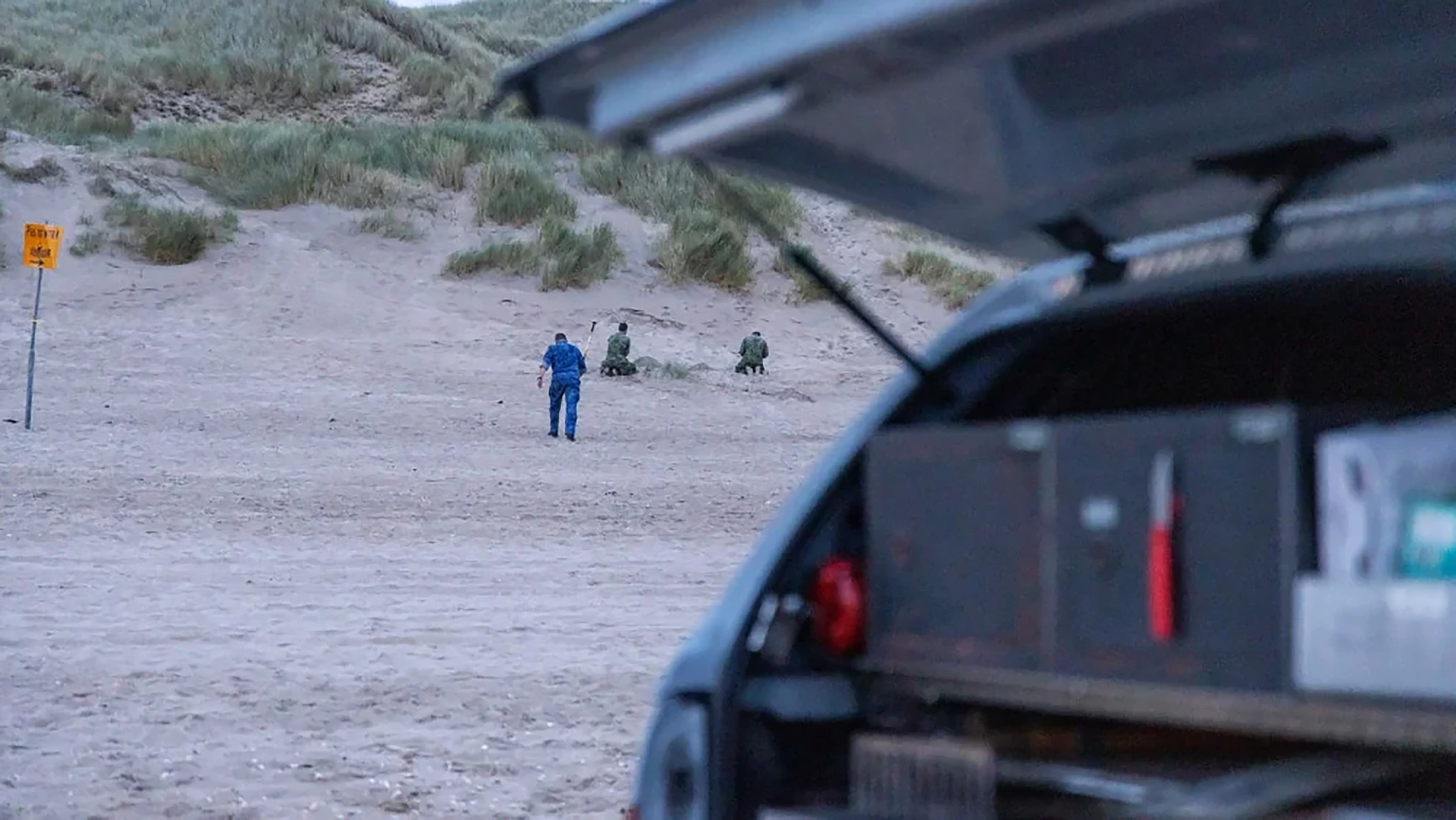 Man loopt over zanderig duingebied naar twee knielende militairen, met open achterklep van auto op de voorgrond