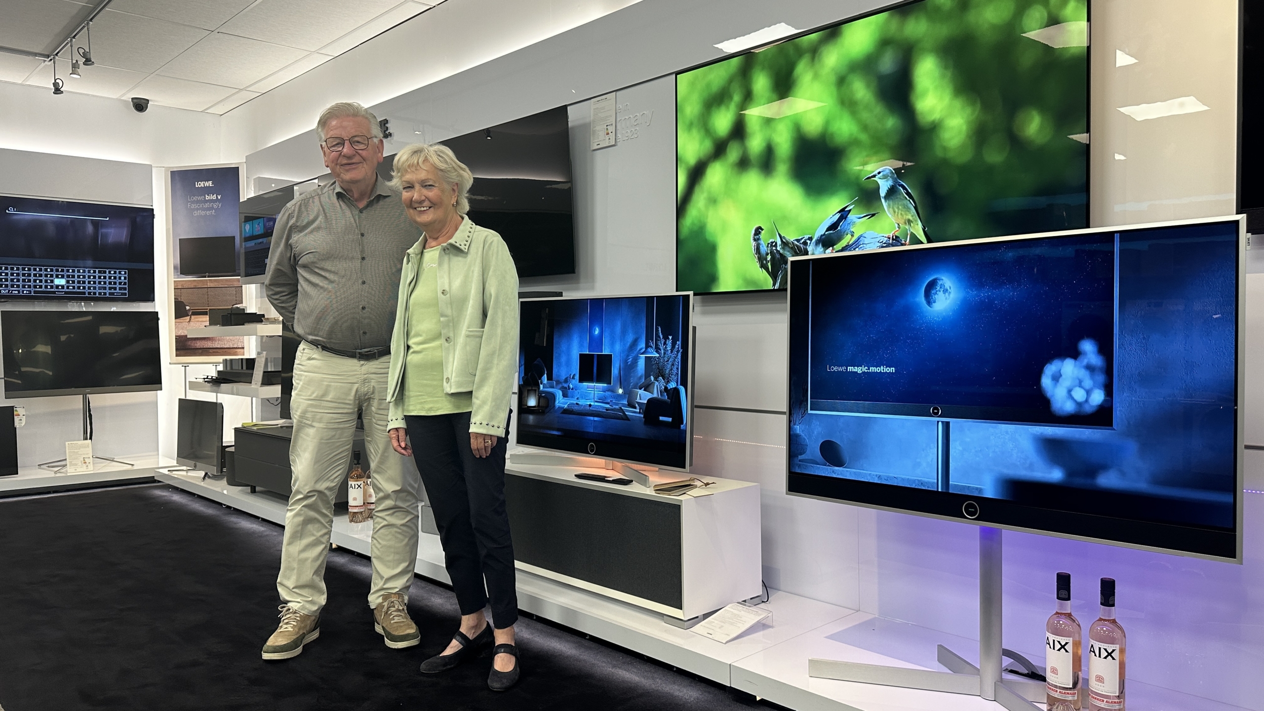 Ouder stel poseert in een tv-winkel tussen grote flatscreens met natuurfoto’s en het scherm met tekst Loewe magic.motion