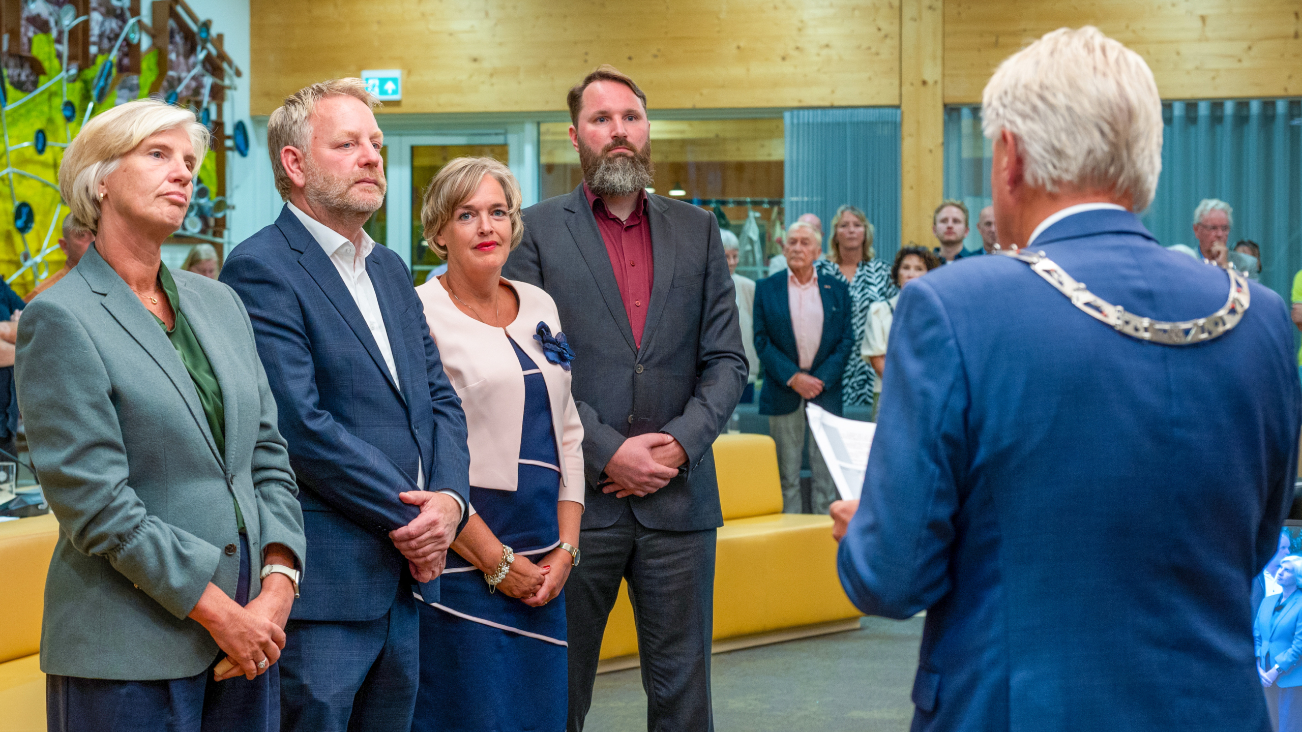 Vier formeel geklede mensen luisteren naar een man met ambtsketen die een toespraak houdt in een zaal