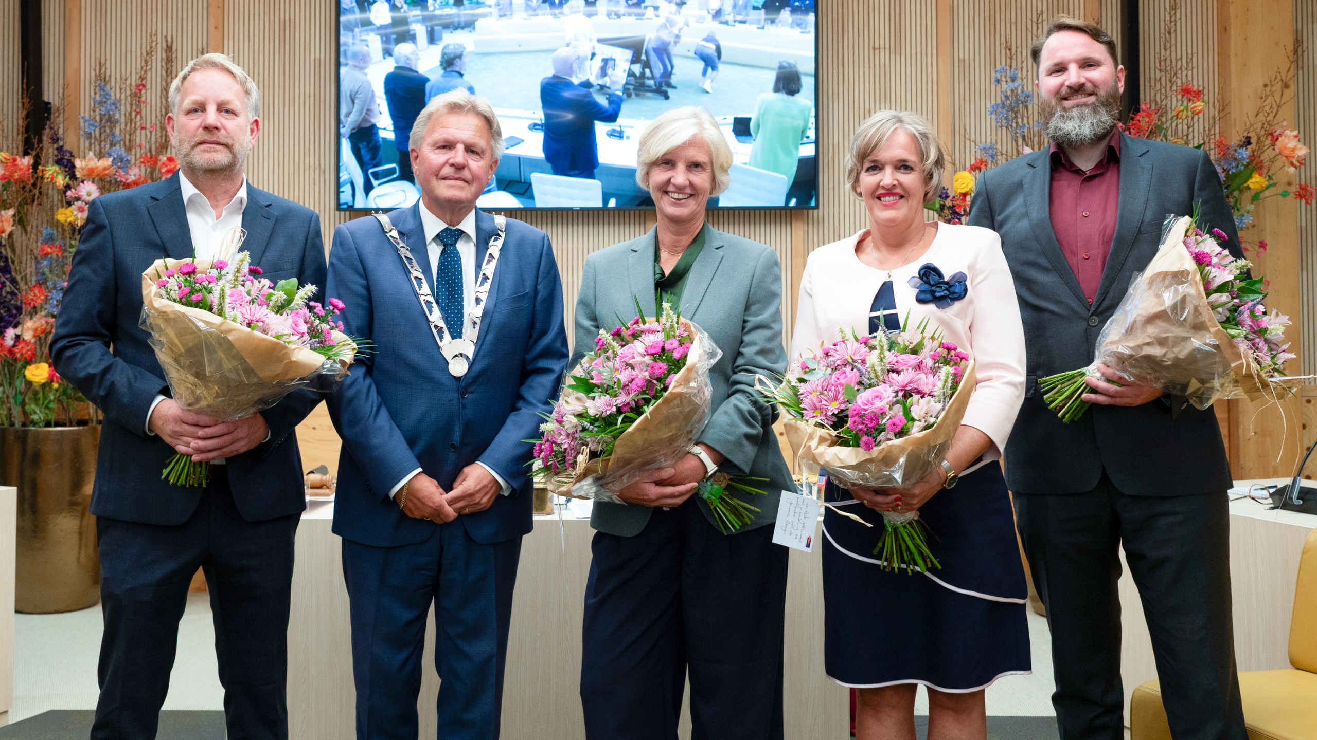Vijf mensen in formele kleding poseren binnen; drie houden grote boeketten vast, met een scherm en bloemen op de achtergrond