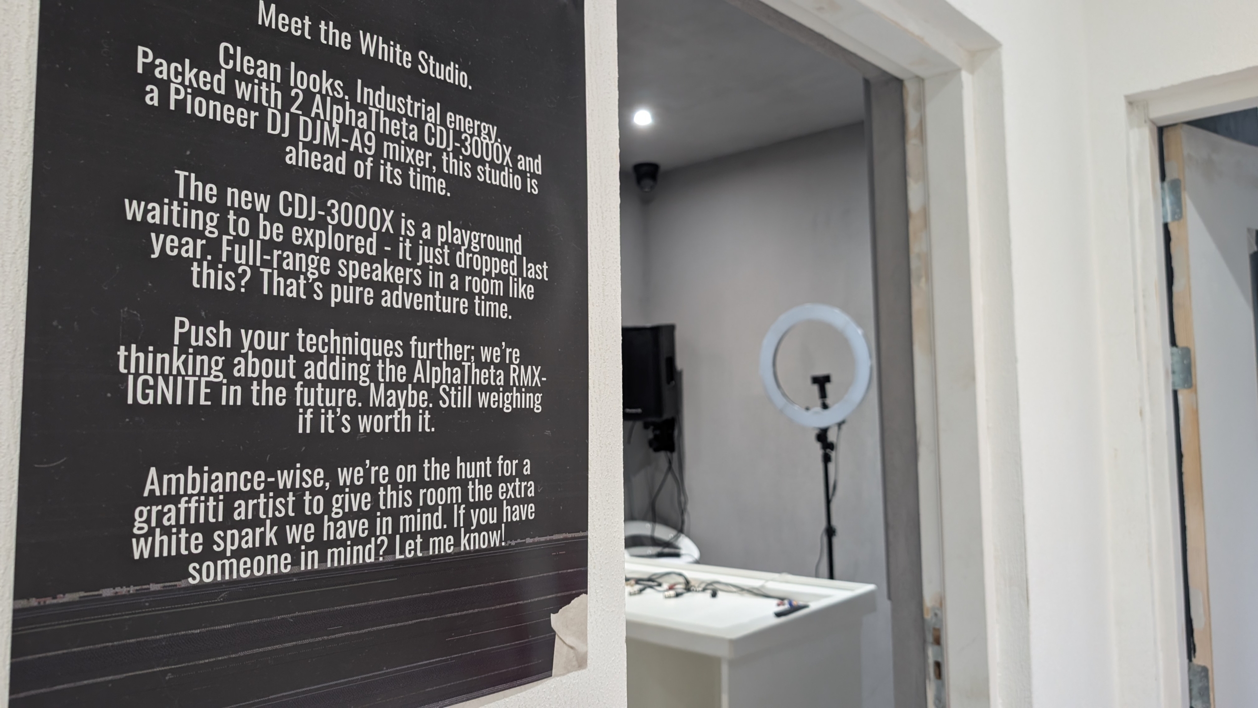 Zwart informatiebord met tekst over “White Studio” naast een open deur naar een witte kamer met ringlamp en apparatuur