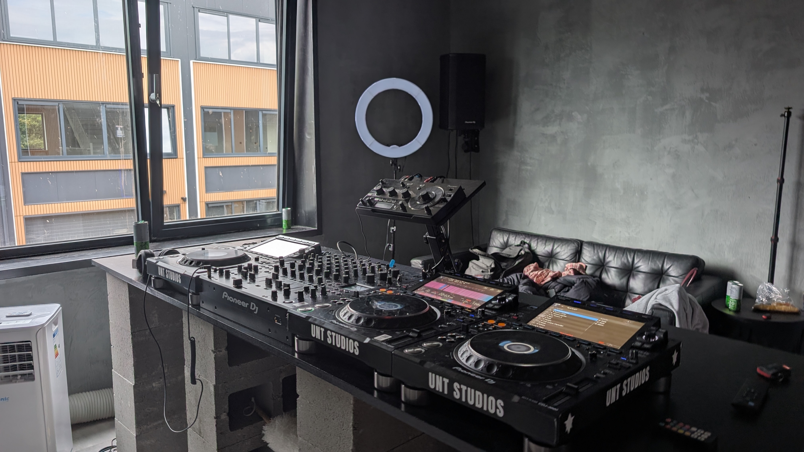 DJ-studio met Pioneer DJ-decks en mengpaneel bij een raam, ringlamp en zwarte bank tegen grijze muur