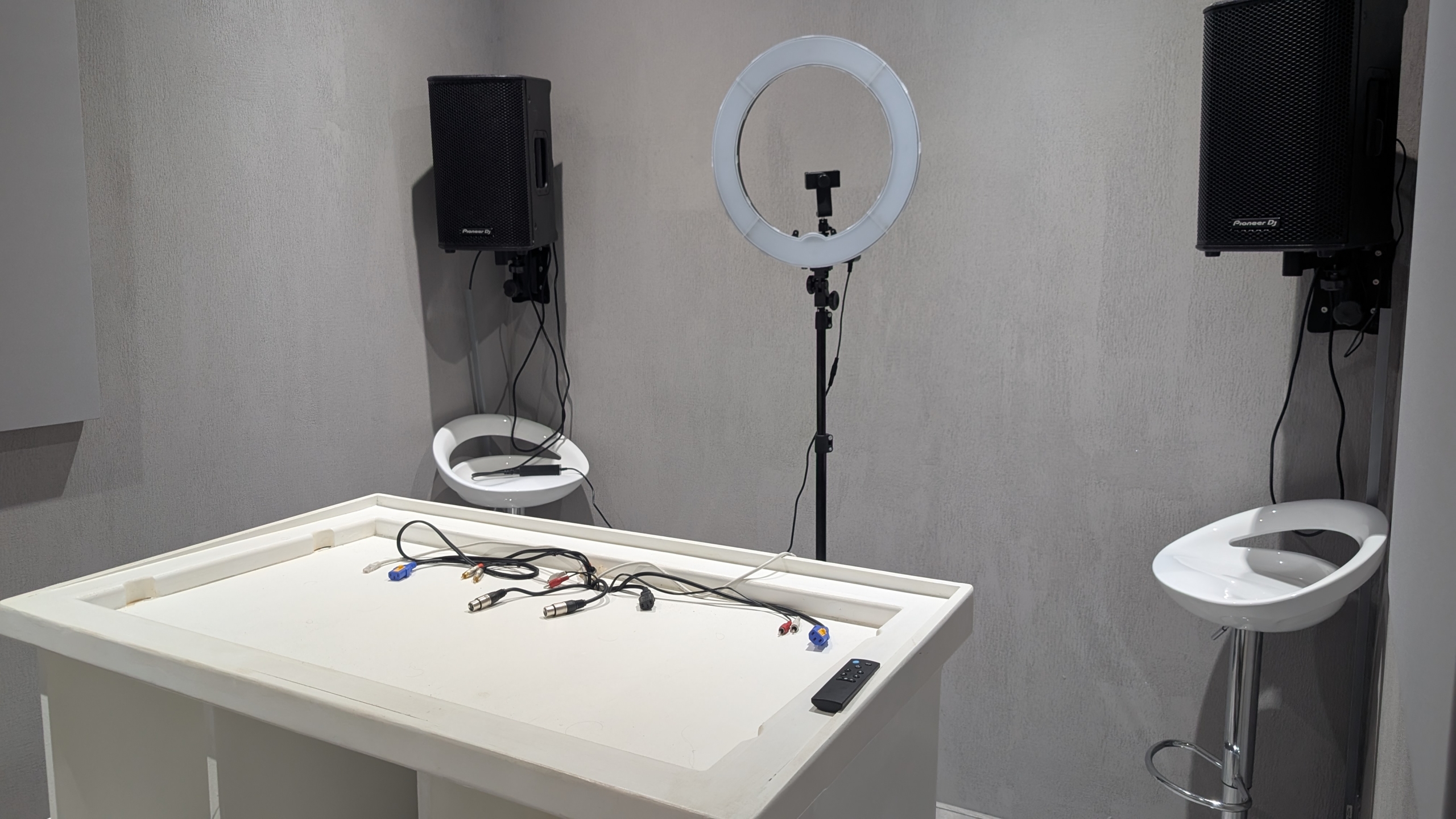 Studiohoek met ringlamp op statief, twee wandgemonteerde speakers, twee krukken en kabels op een witte tafel