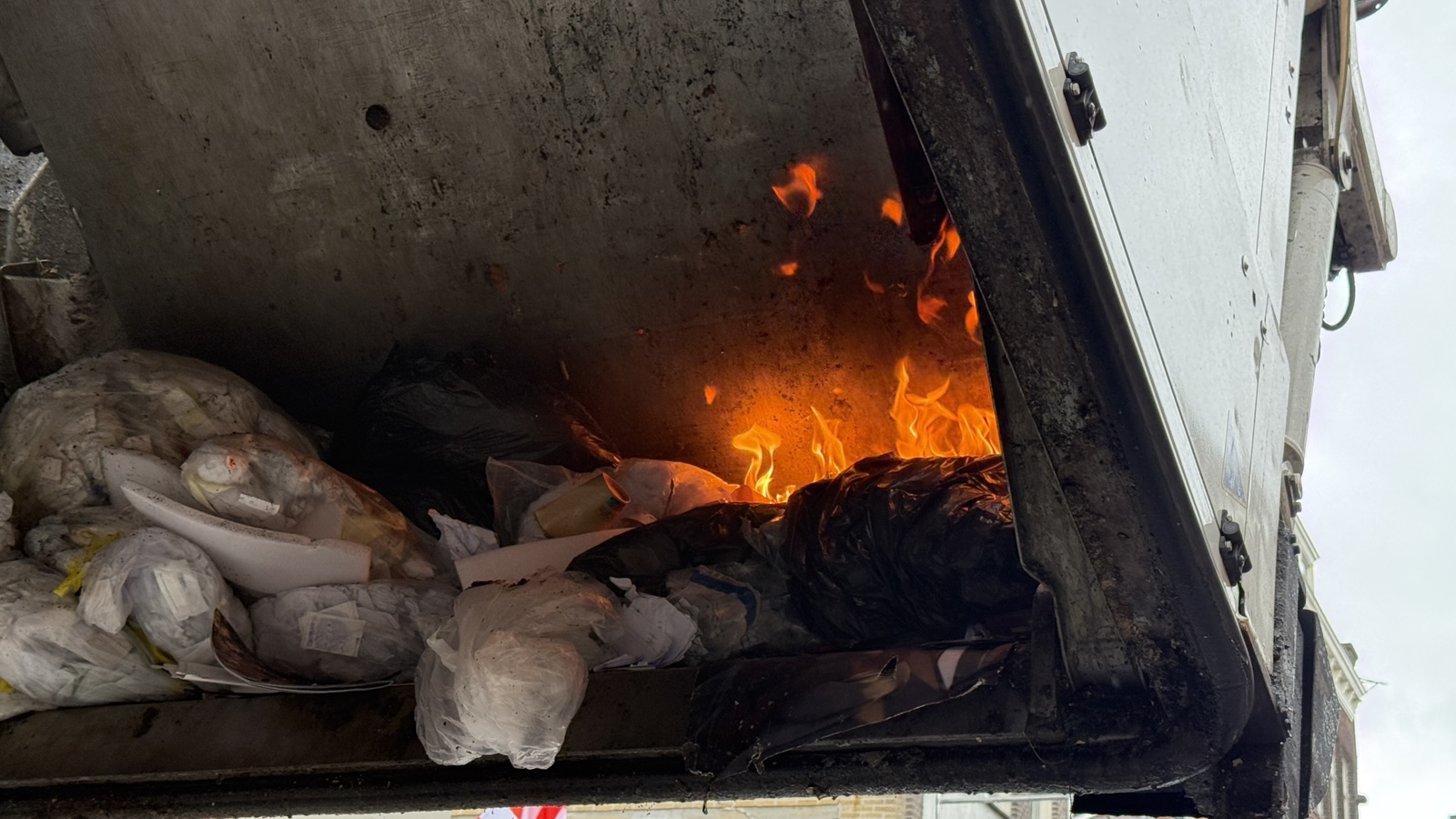 Vlammen in de achterzijde van een vuilniswagen tussen zwarte en witte vuilniszakken en afval
