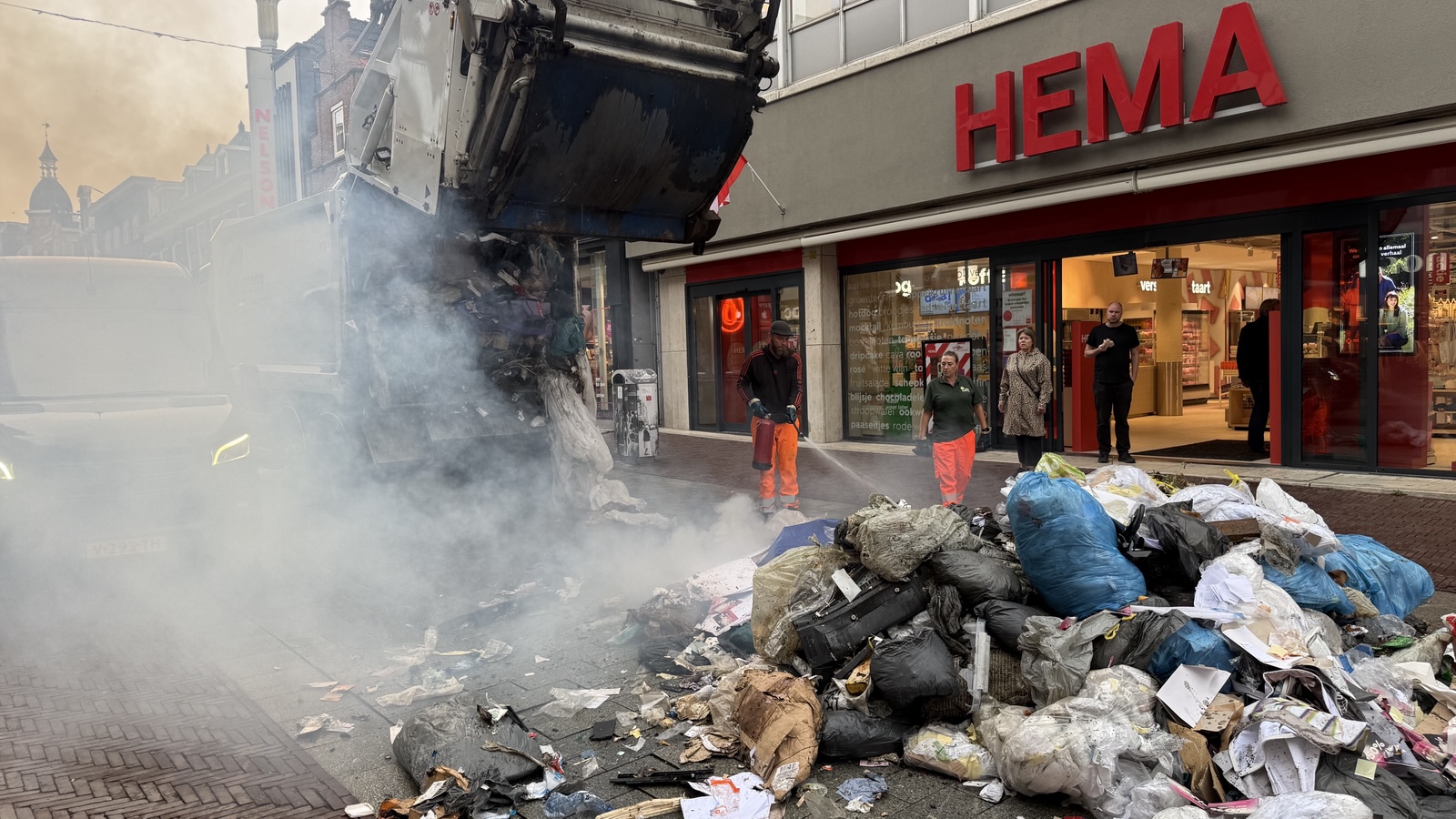 Vuilniswagen lost rokende afvalberg voor de HEMA, terwijl twee medewerkers blussen en omstanders bij de ingang staan