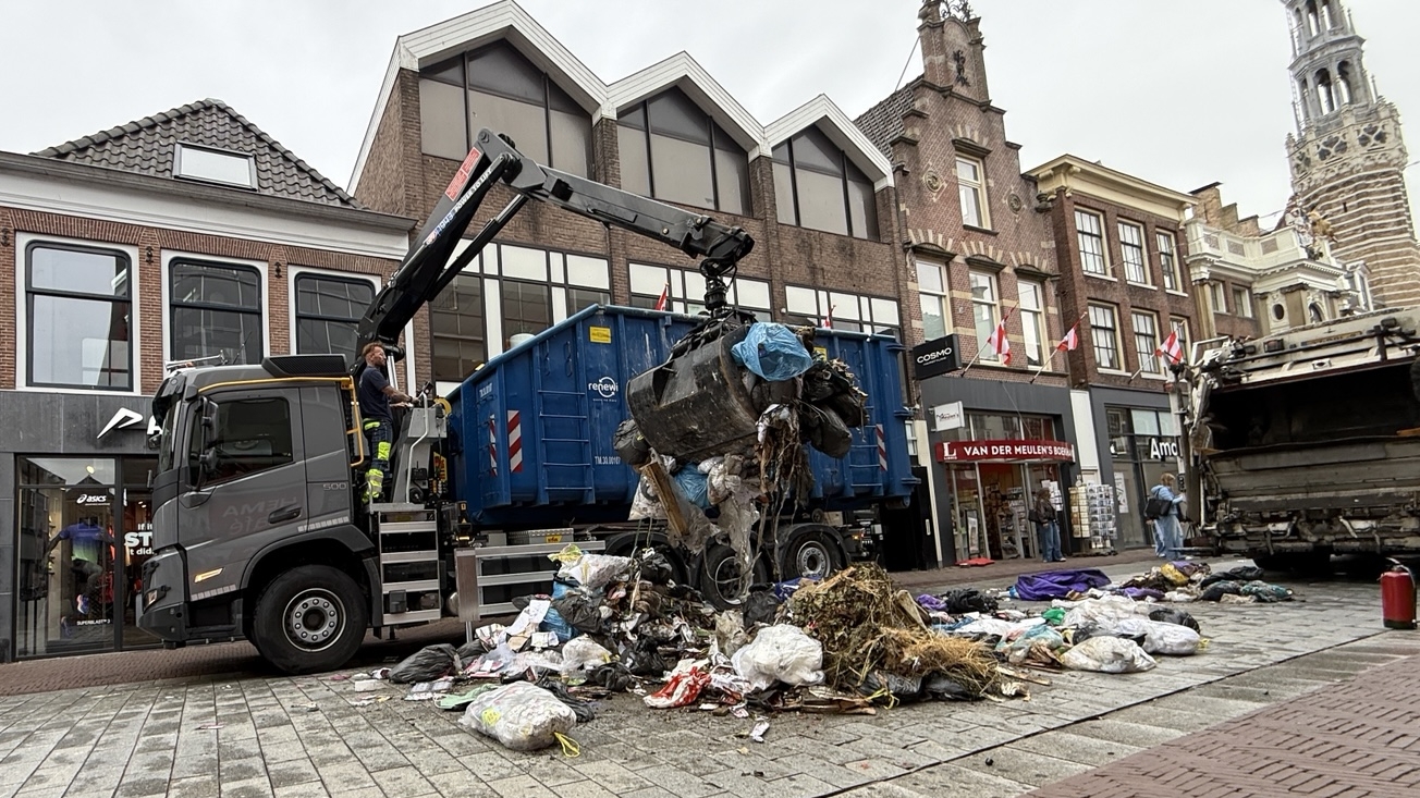 Vuilniswagen met kraan laadt een grote hoop afval van het plein, met winkels en oude gevels op de achtergrond