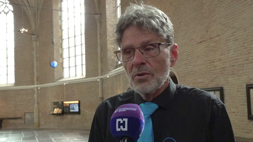 Man met bril en grijze baard wordt geïnterviewd met microfoon in een grote kerkruimte met hoge ramen