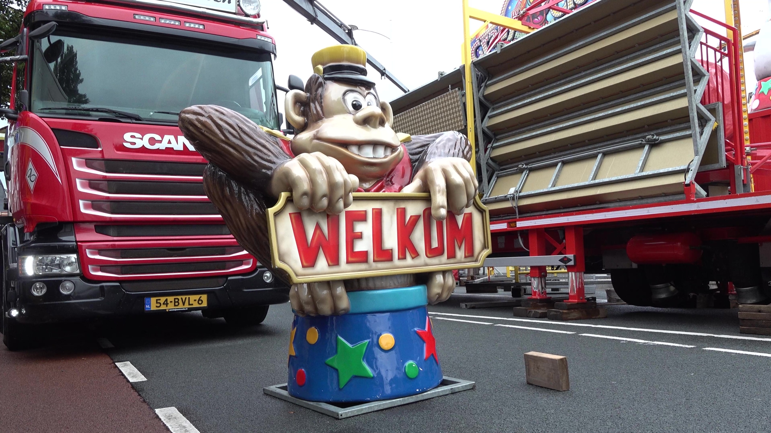Apenfiguur met bord ‘WELKUM’ op straat, voor een rode vrachtwagen en een kermiswagen met uitklapbare tribune