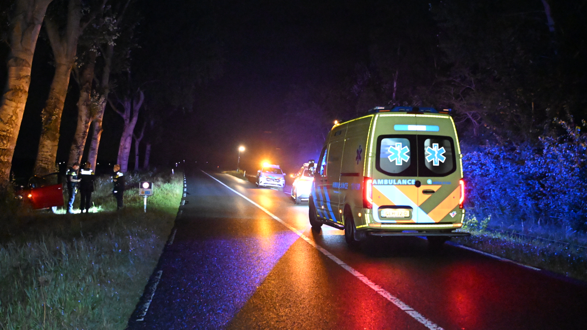 Ambulance en politievoertuigen met zwaailichten op een donkere weg, hulpverleners bij een auto in de berm