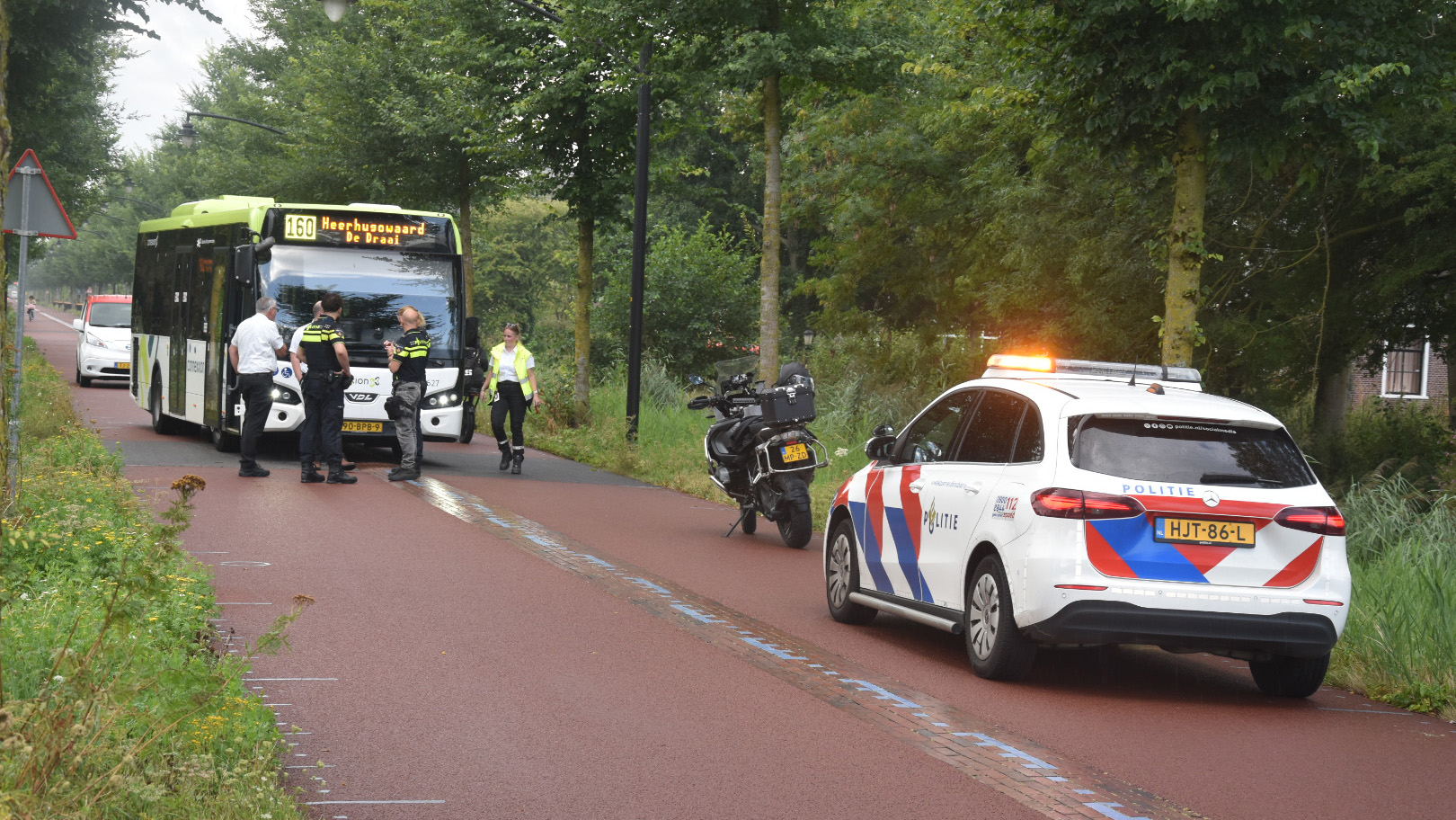 Politieauto met zwaailicht naast stilstaande lijnbus 160; agenten en bestuurder praten op een bosrijke weg