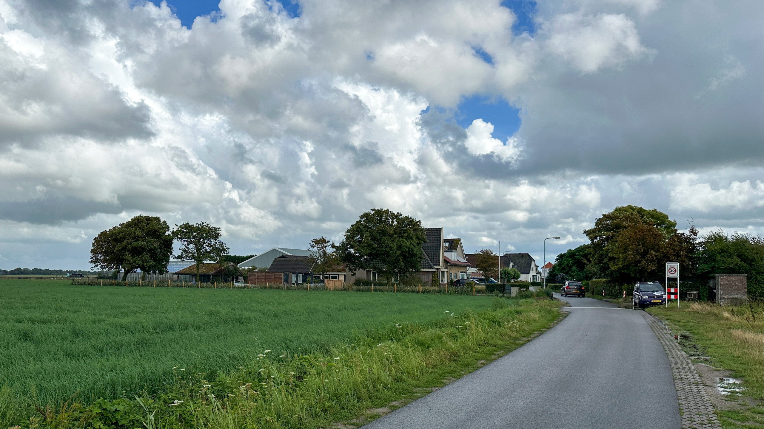 Landweg langs groen weiland met boerderijen en geparkeerde auto’s, onder een lucht met grote stapelwolken
