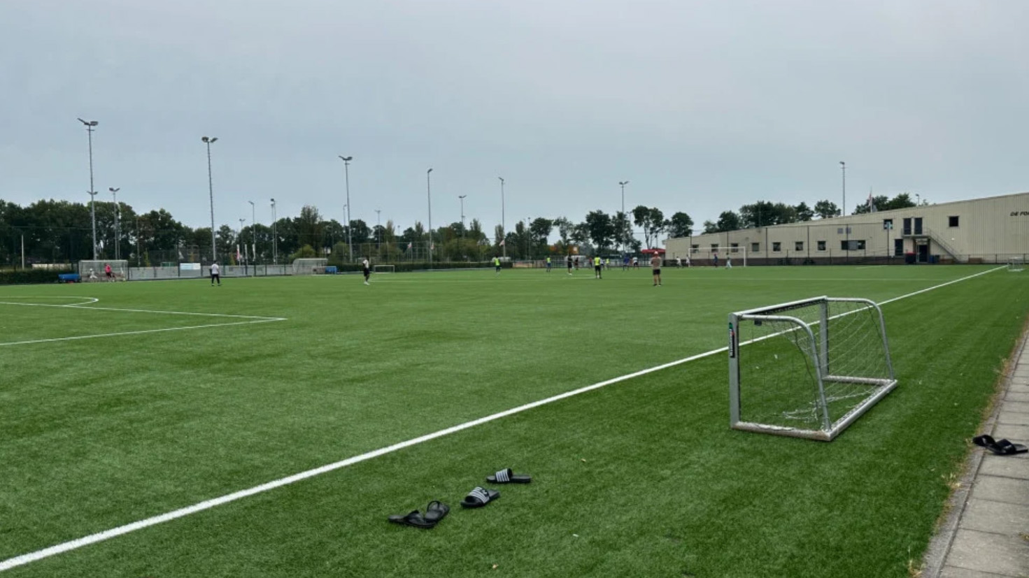 Voetbalveld met spelers in de verte, klein doel aan de zijlijn en slippers op het kunstgras onder grijze lucht