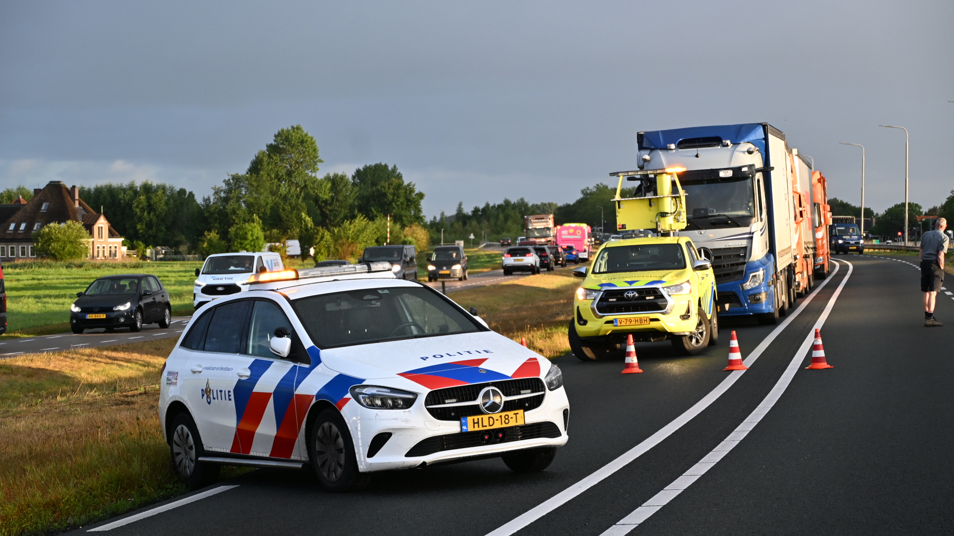 Politieauto met zwaailicht blokkeert rijstrook met pionnen; gele hulpdienstauto en vrachtwagens staan stil in file