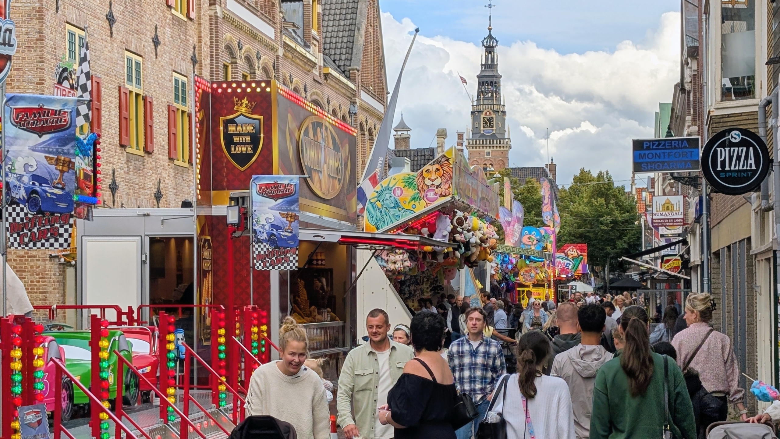 Drukke kermisstraat met attracties en kraampjes, veel bezoekers en een klokkentoren op de achtergrond