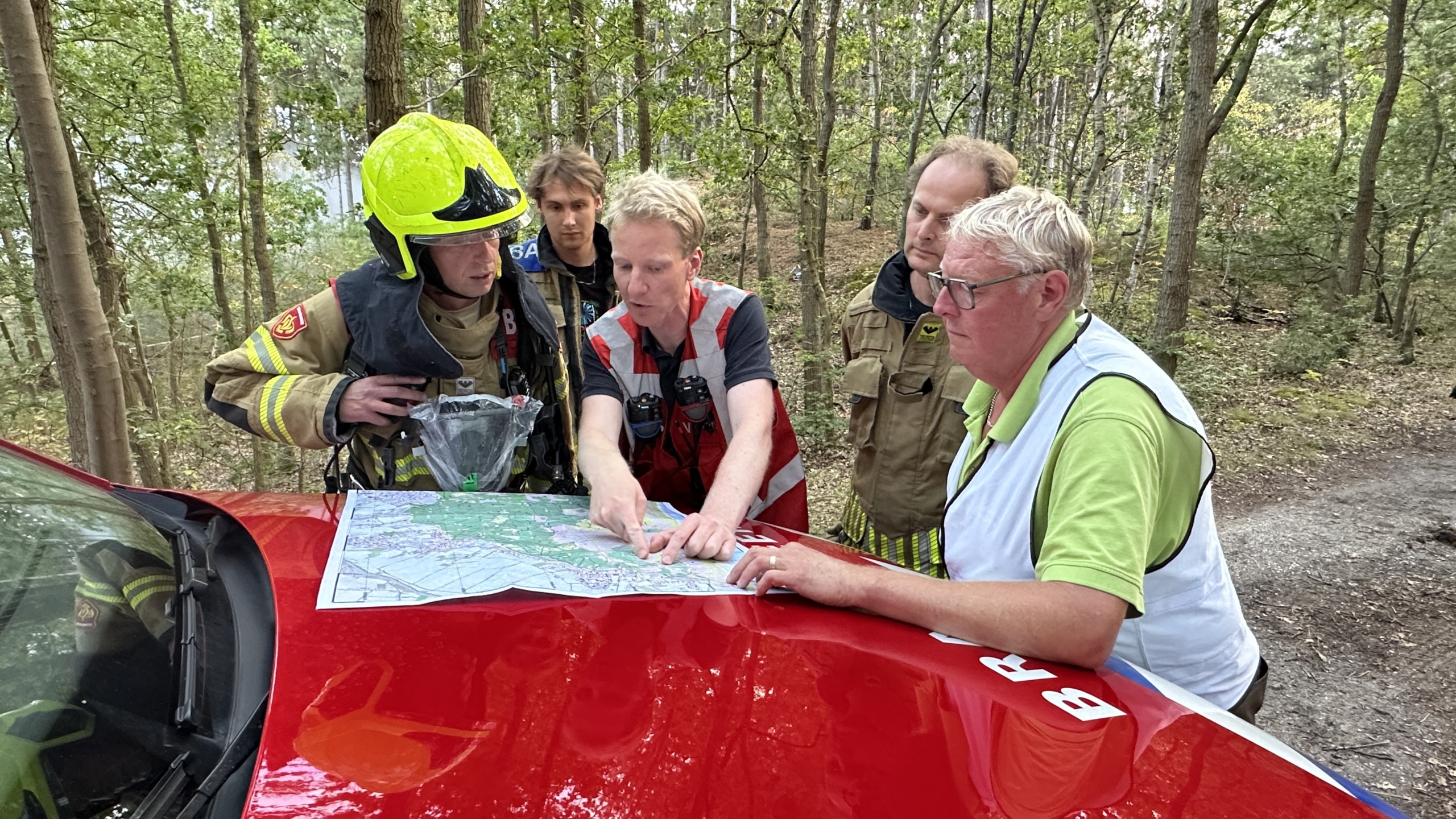 Brandweer en hulpdiensten bespreken een kaart op de motorkap van een rode auto in een bosgebied