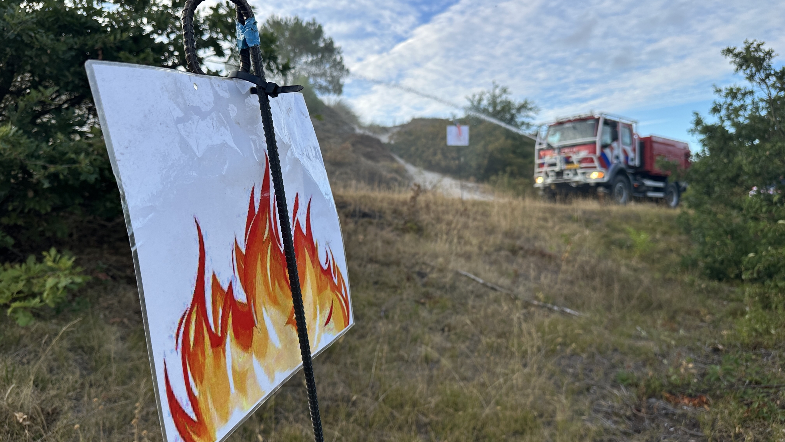 Bord met vlammenpictogram op de voorgrond, met op de achtergrond een brandweerwagen die water spuit over een heuvel